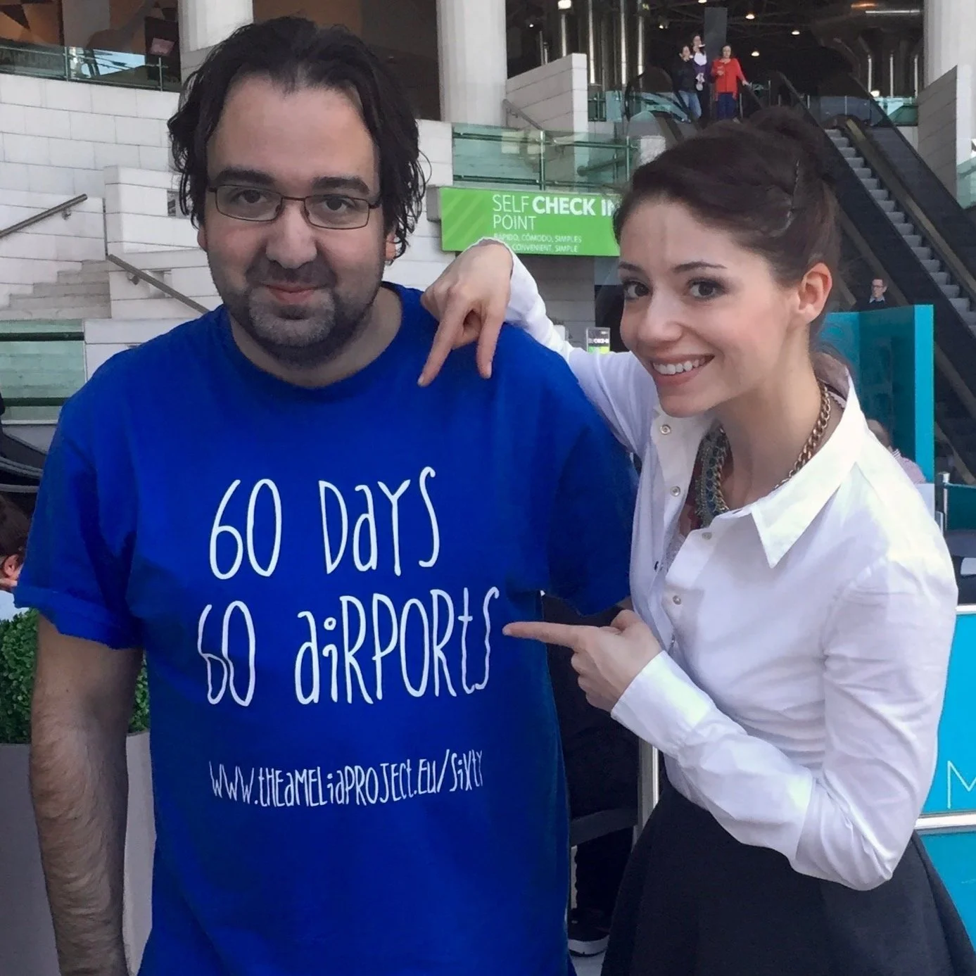 A man and a woman standing together at an indoor event. The man is wearing glasses and a blue T-shirt that reads '60 DAYS 60 AIRPORTS' and a website address. The woman is smiling, pointing at the man's shirt, and has her arm around his shoulder. They are in a modern public space with escalators and glass railings in the background, and other people are visible in the distance.