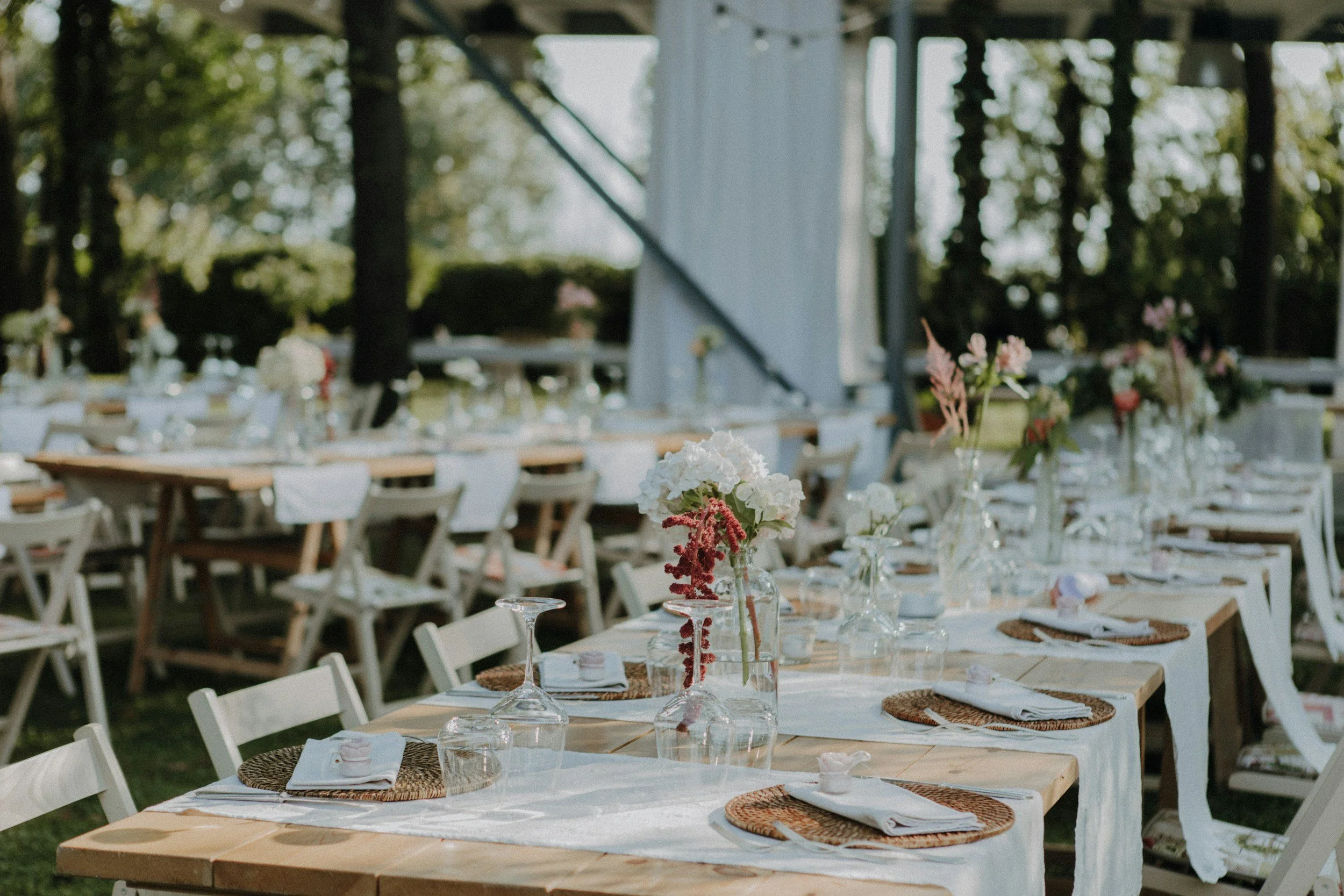 Outdoor wedding reception tables with white tablecloths, floral centerpieces, glassware, and place settings in a wooded area with sunlight filtering through trees.