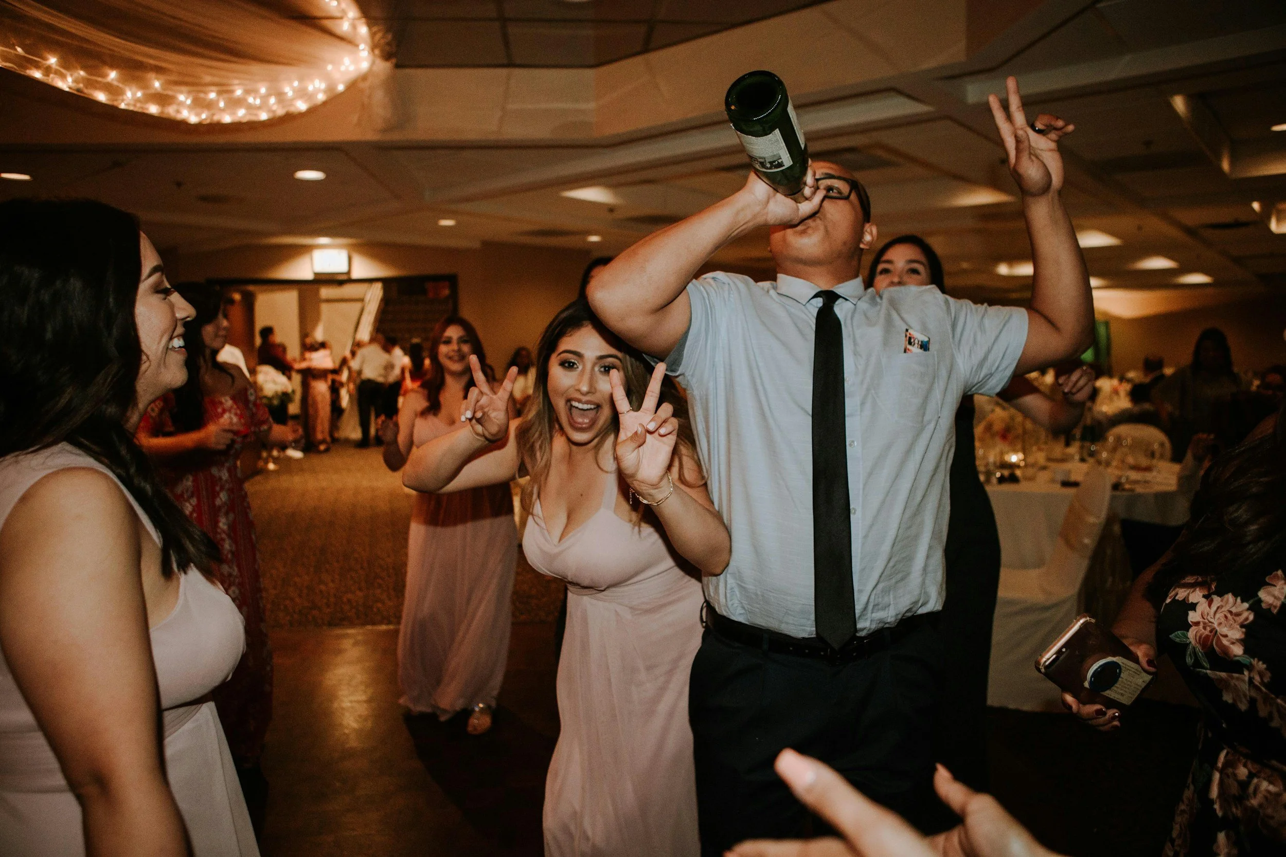 People celebrating at a party, with one man drinking from a bottle and others smiling and making peace signs.