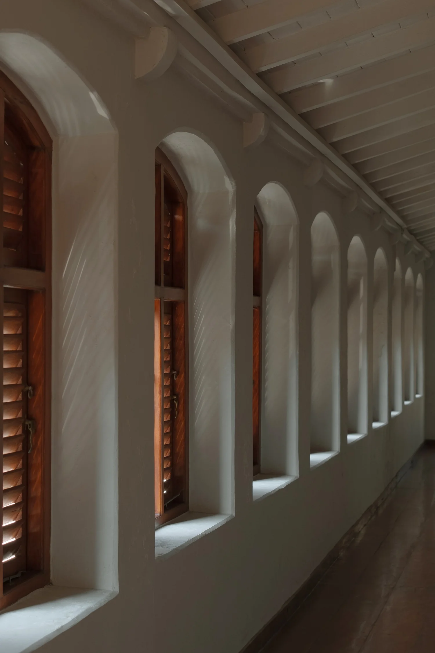 A long whitewashed gallery lit by a row of arched windows with wooden shutters