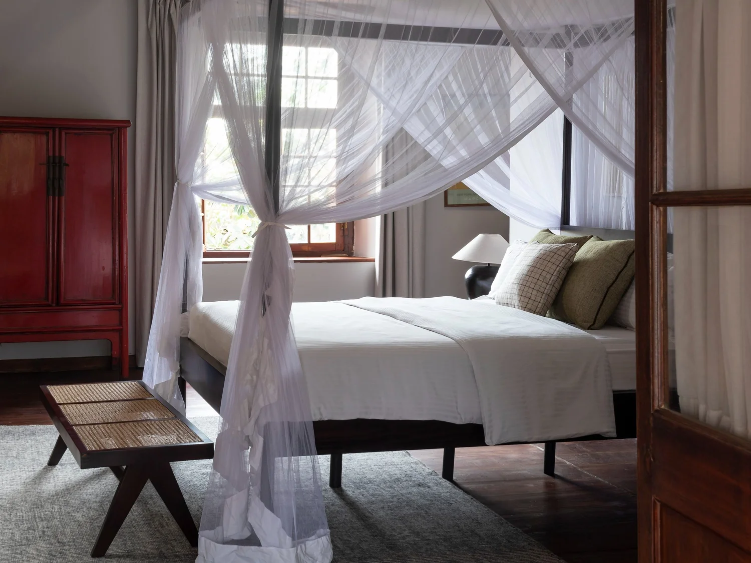A four-poster bed hung with white netting, beside a red lacquered cabinet and a low cane bench