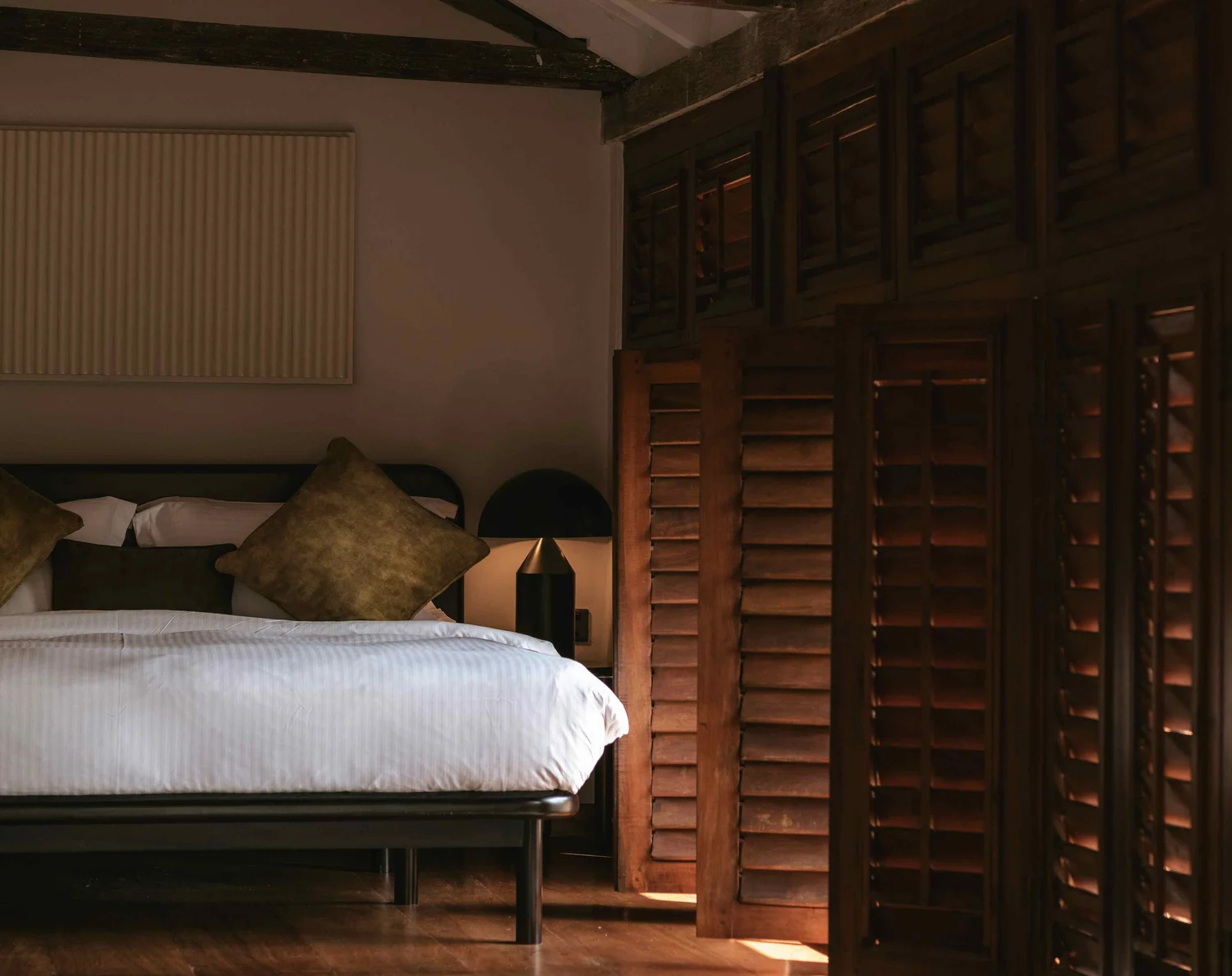 A bedroom under a pitched timber roof, with a white bed and a wall of dark wooden louvred shutters