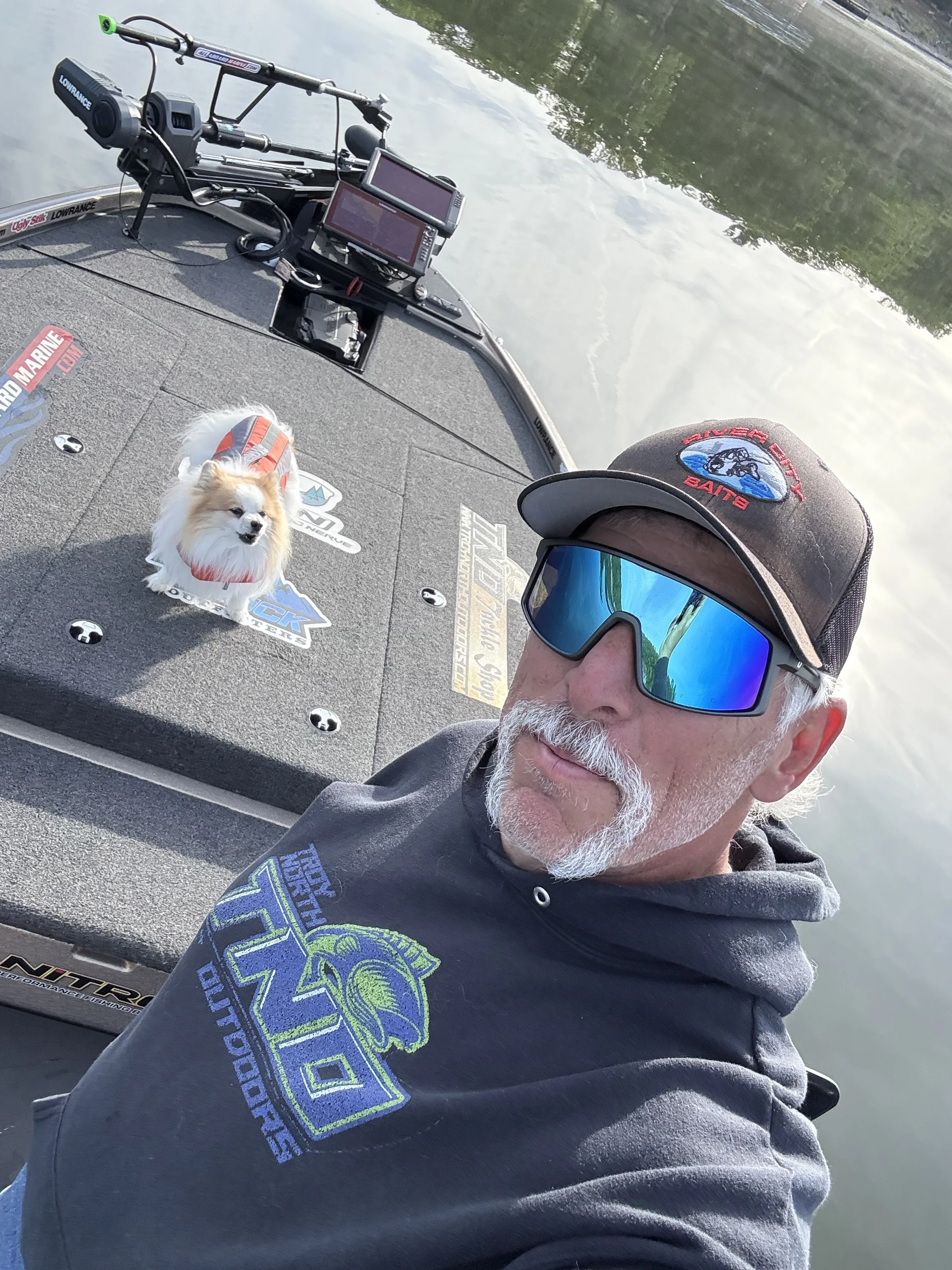 A man taking a selfie on a boat, wearing a cap, sunglasses, and a hoodie. A small fluffy dog is sitting on the boat deck nearby, wearing a red and gray life vest. The boat has various fishing and electronic equipment, and the water and trees are visible in the background.