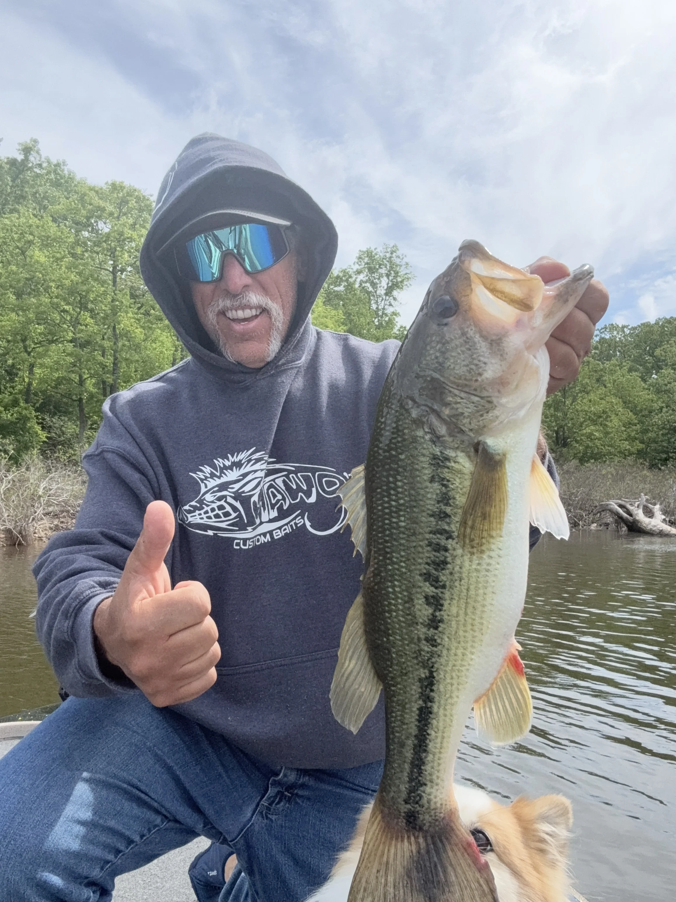 A man wearing a hoodie, sunglasses, and a T-shirt with a logo, holding a large bass fish with one hand, giving a thumbs-up while sitting outdoors on a boat or dock near a river or lake, with trees and a partly cloudy sky in the background.