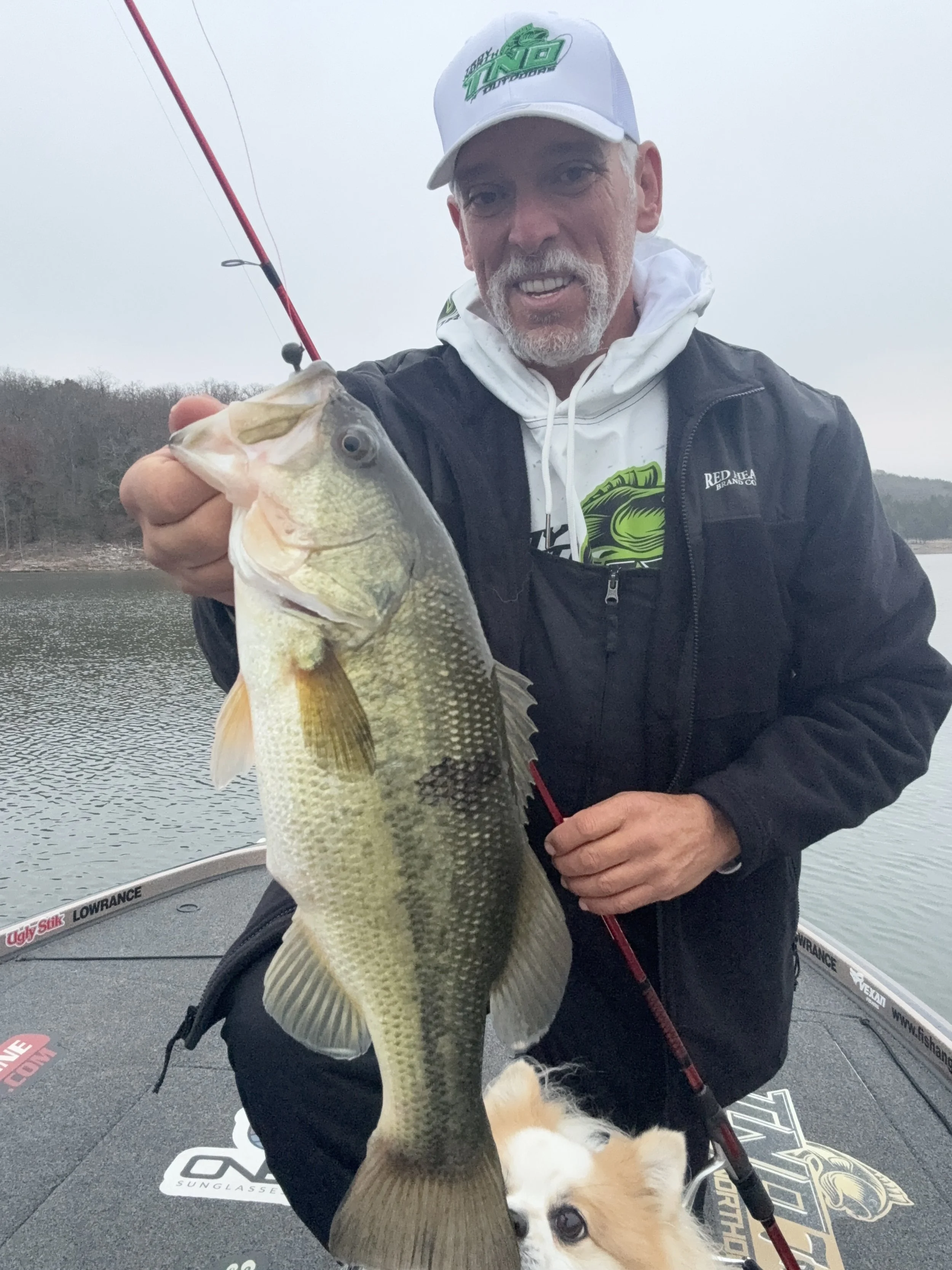 Troy holding a largemouth bass fish on a boat with water and trees in the background, wearing a white cap and black jacket, with Winston his dog at his feet.