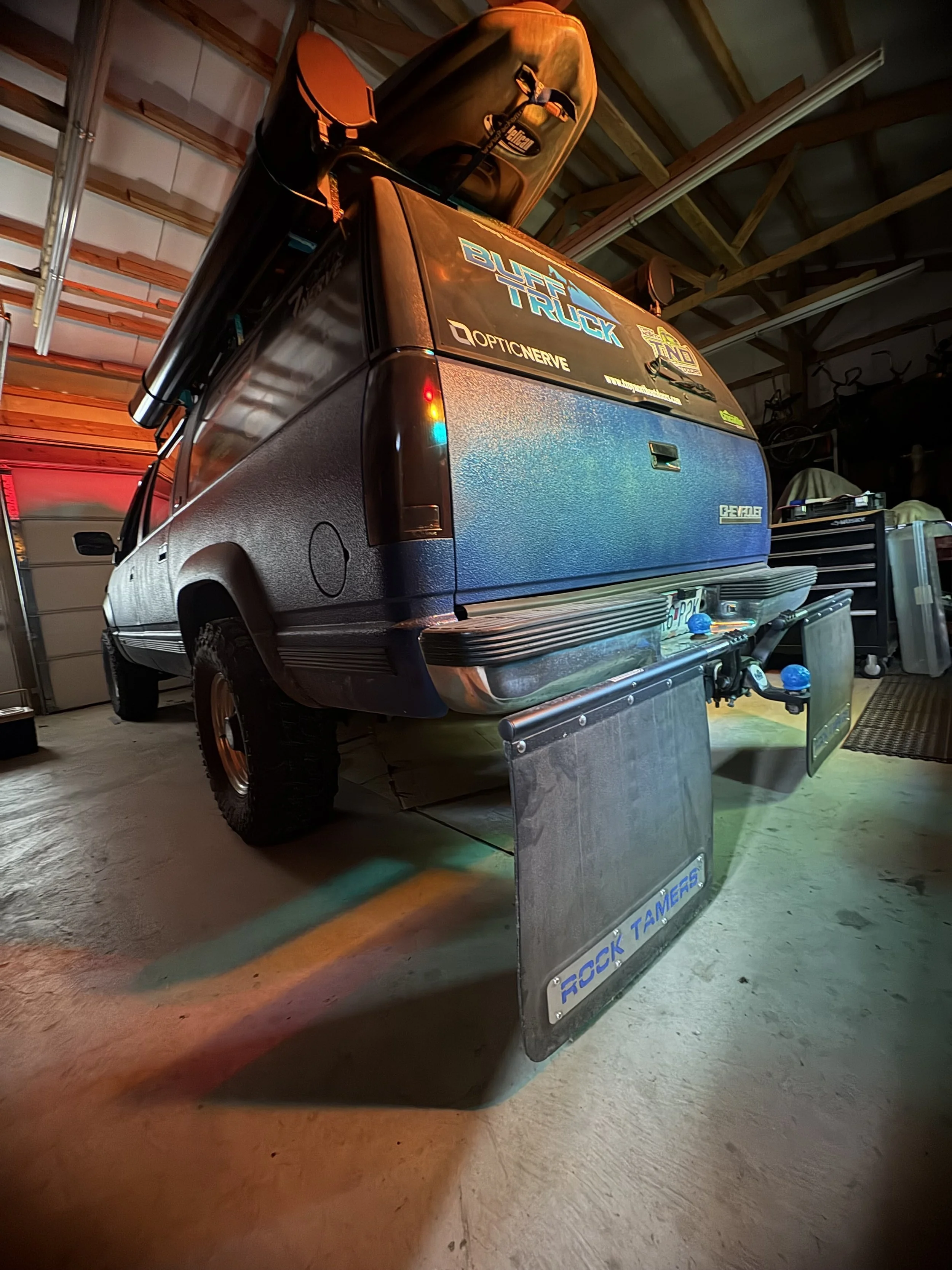A black pickup truck parked inside a garage with various stickers on the back, including 'BUFF TRUCK' and 'OPTICNERVE,' with a large metal toolbox labeled 'ROCK SHOCK' placed on the floor in front of the truck.