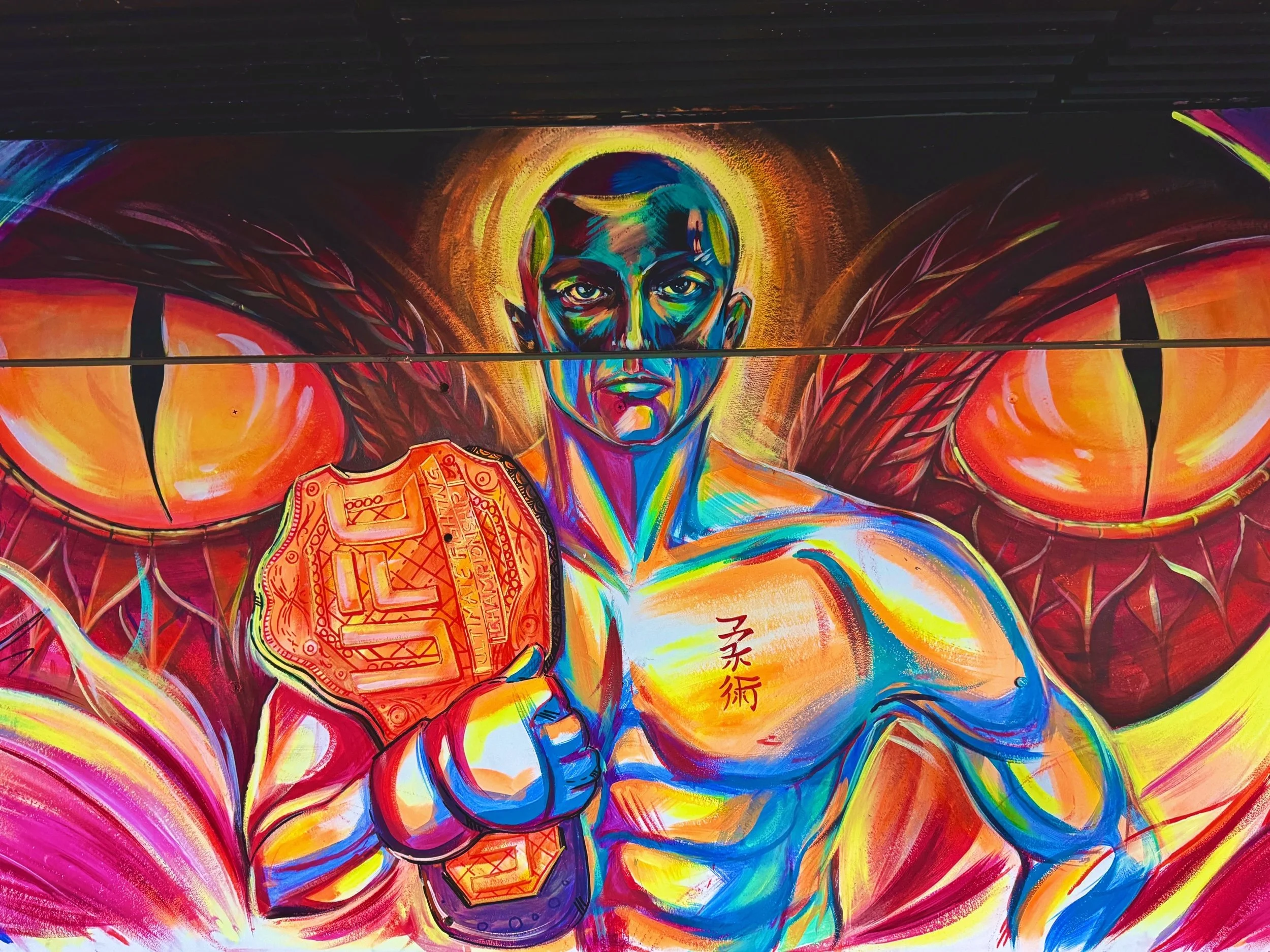 Colorful graffiti mural featuring a portrait of a person with a shaved head, bright expressive eyes, and raised fists. The mural incorporates abstract and vibrant elements, including a stylized badge or emblem on one hand and Asian characters on the 