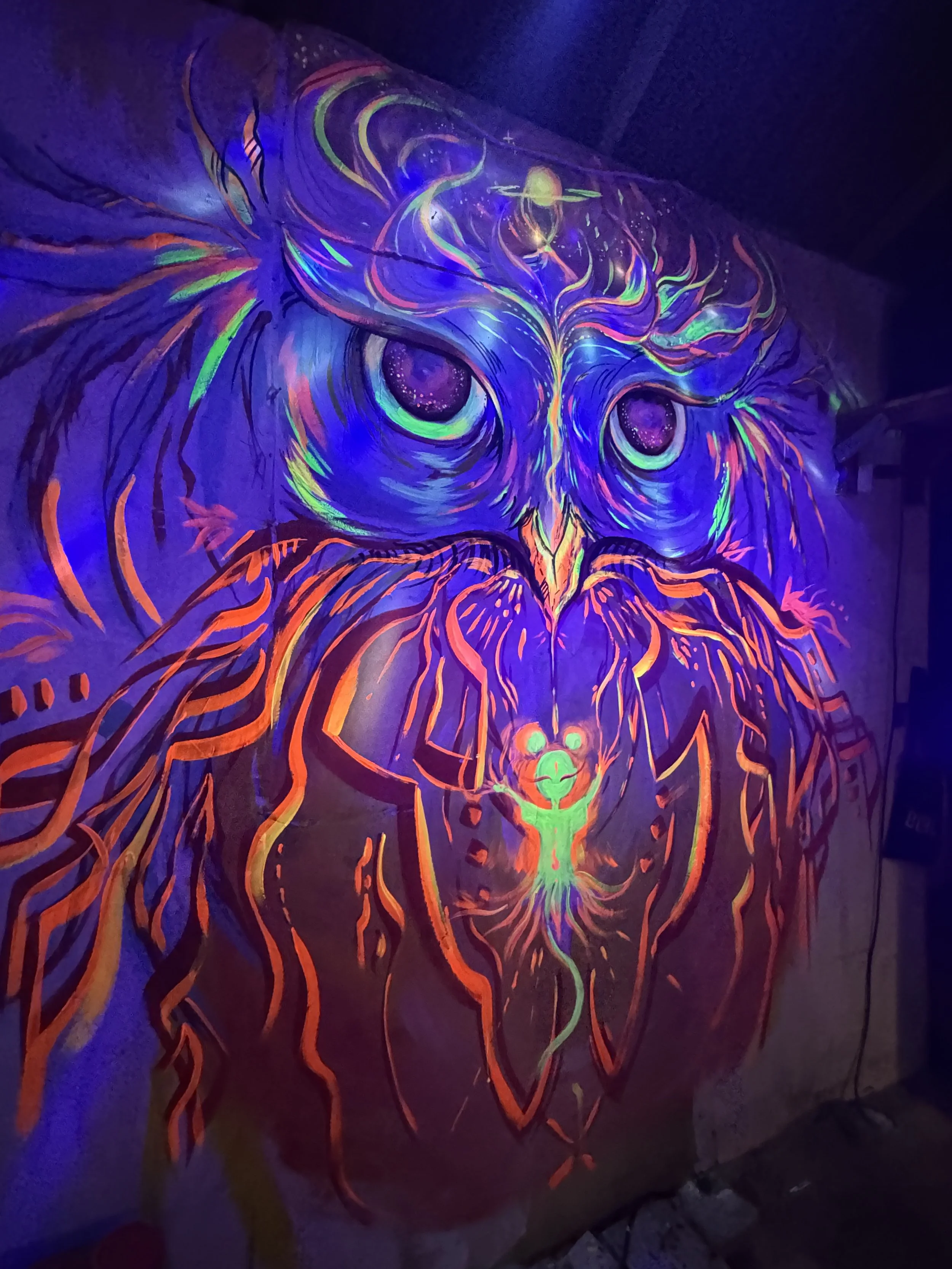 Vibrant street art mural of an owl with glowing eyes, surrounded by swirling patterns and orange flames, illuminated with blacklight effects.