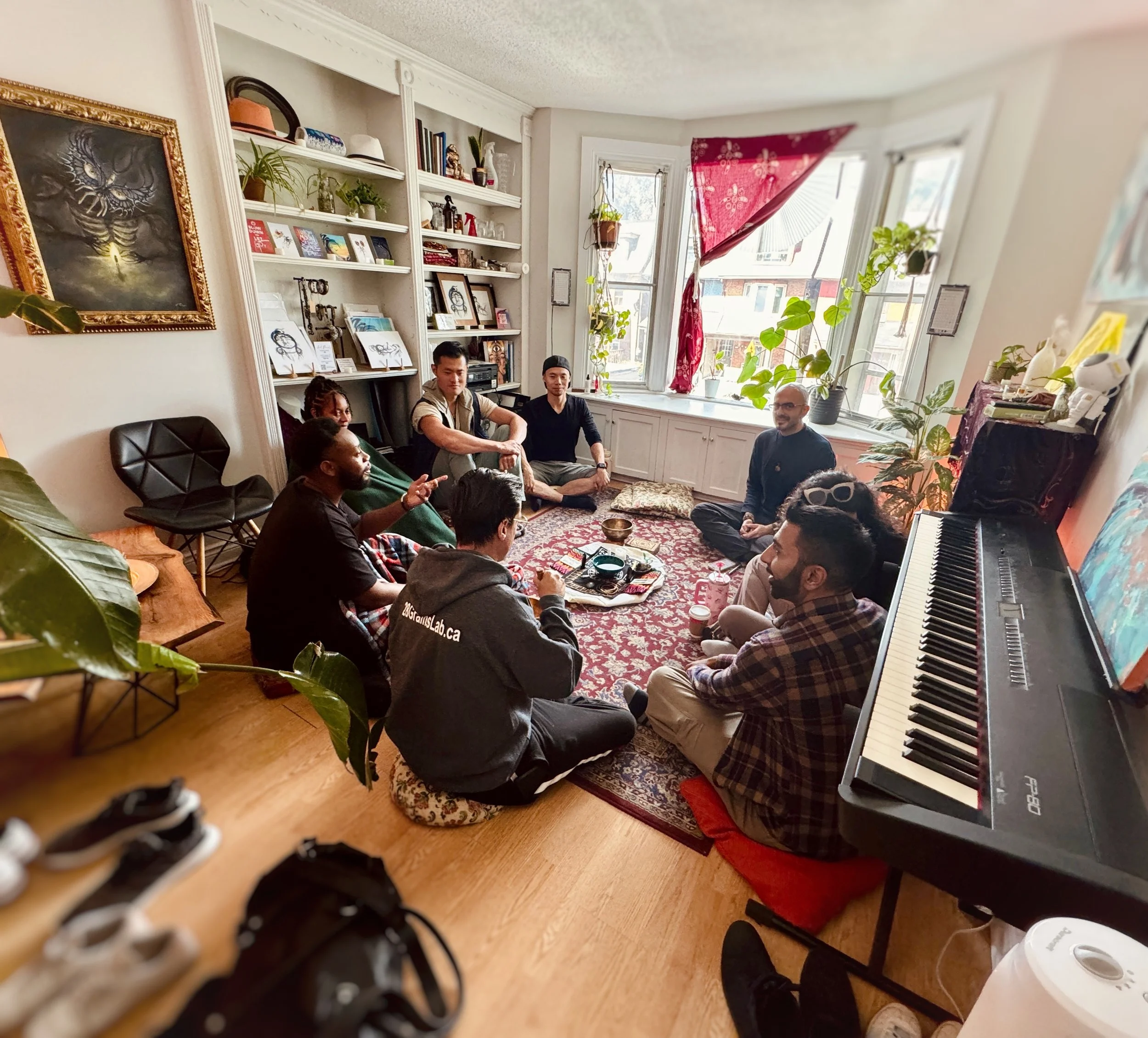 Group of diverse people sitting on the floor in a cozy living room with a kitchen piano, bookshelf, plants, and large windows.