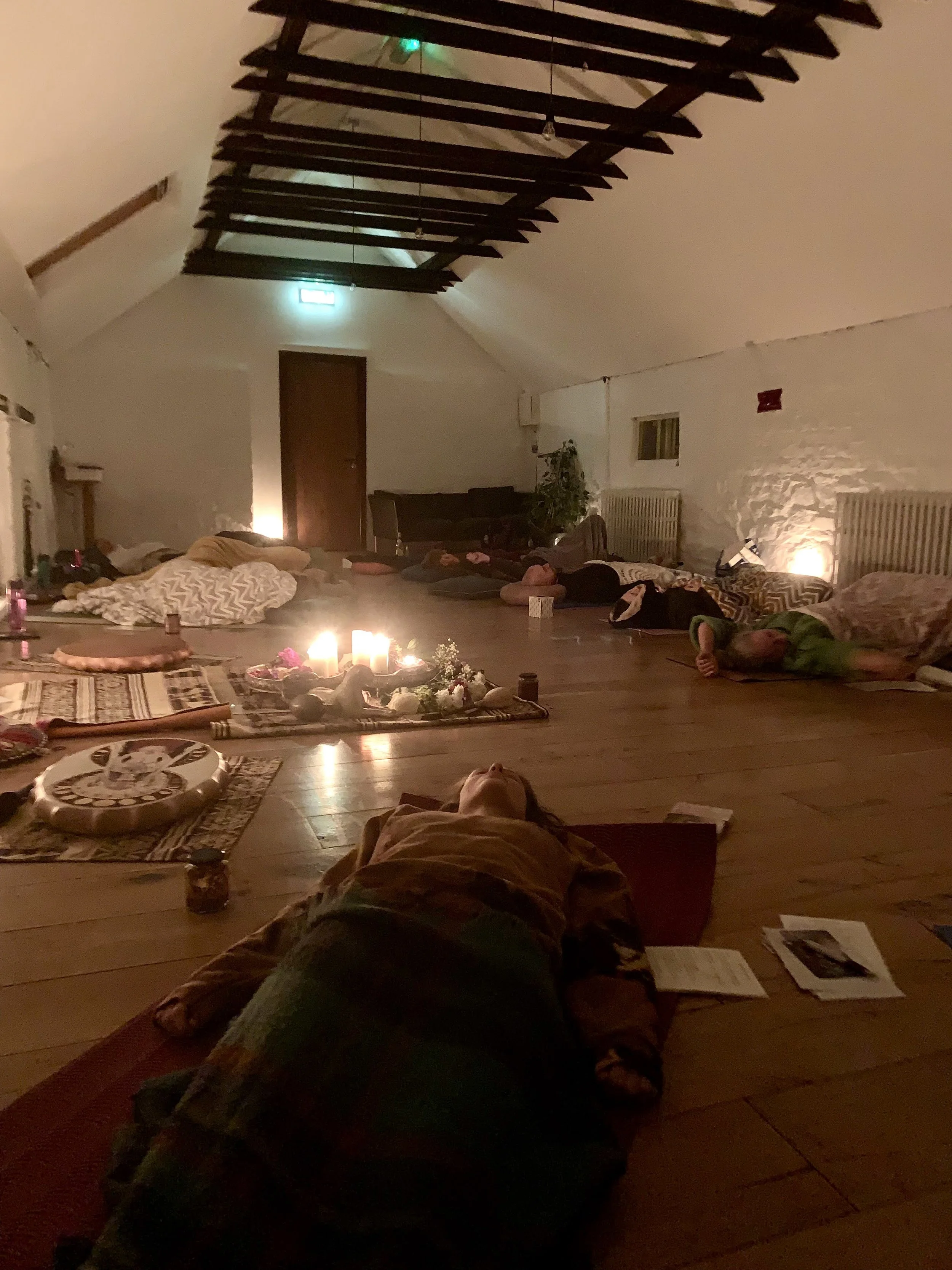 People lying or sitting on the floor of a dimly lit room with candles, pillows, and mats, likely participating in a meditation or yoga session. The room has a high ceiling with wooden beams and soft lighting.