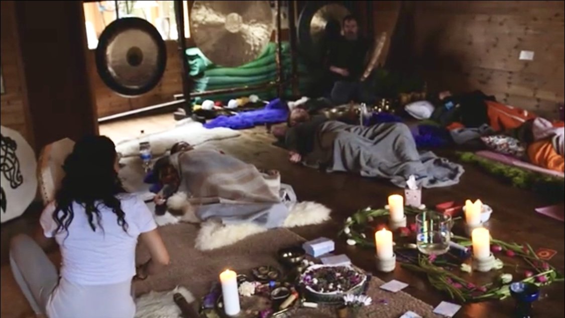 Group of people participating in a sound healing or meditation session, lying on the floor with candles, flowers, and healing objects arranged around them in a cozy wooden room.