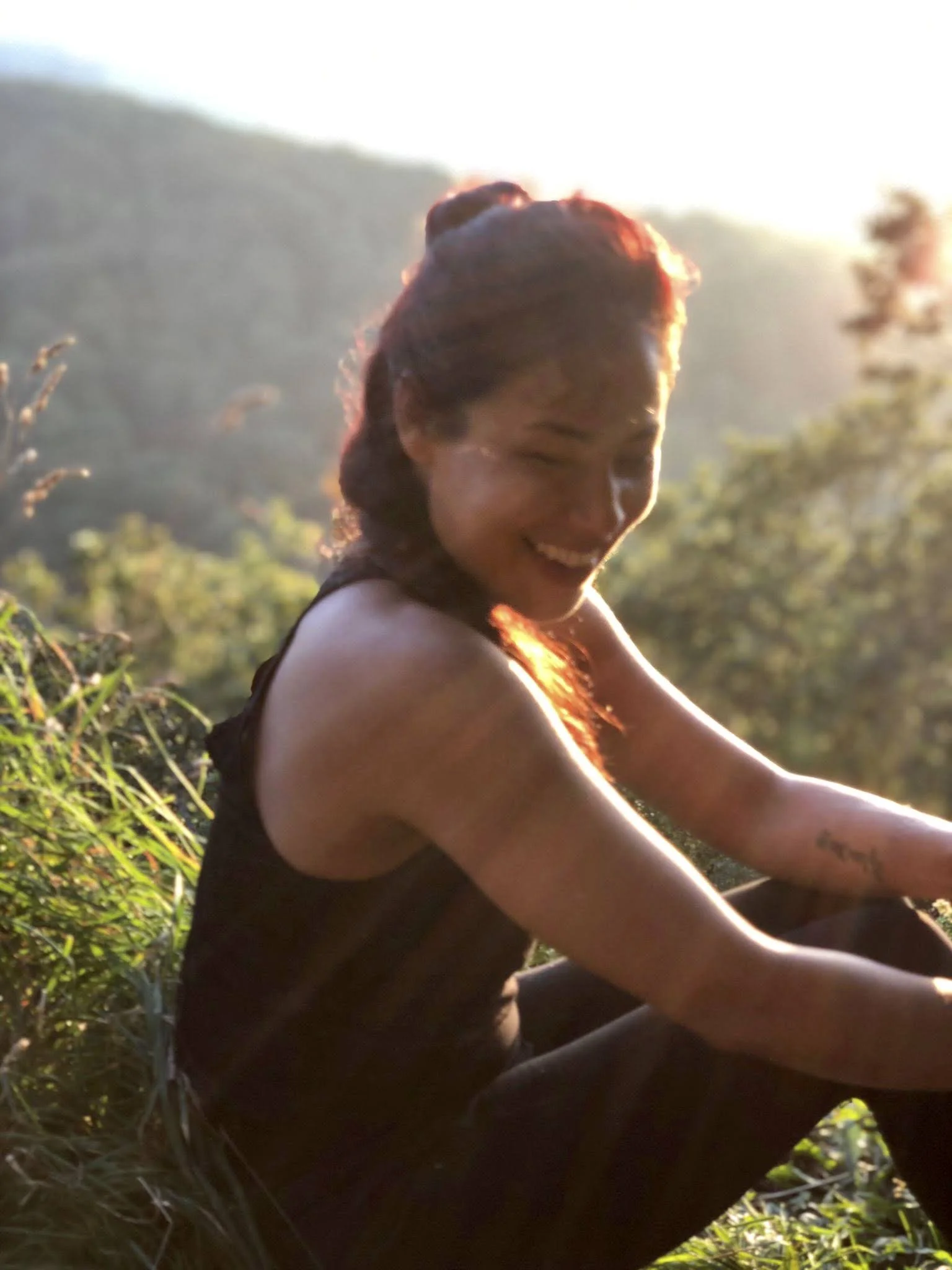 Girl with curly hair smiling and squinting in the sunlight, sitting outdoors among green plants, with a mountain in the background.