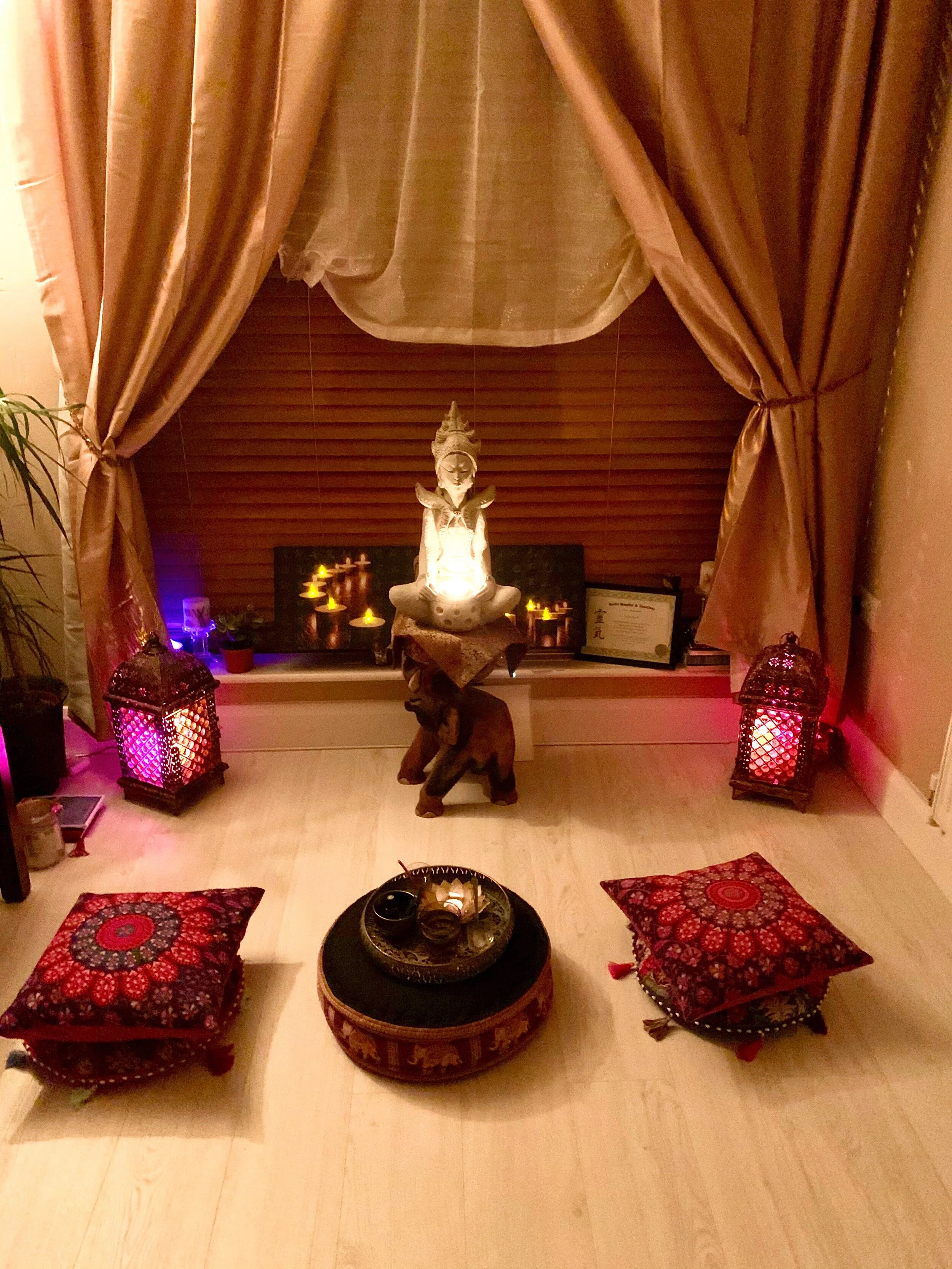 A cozy meditation space with a Buddha statue in the center, surrounded by lit candles, draped curtains, and four red patterned cushions on the light wooden floor.
