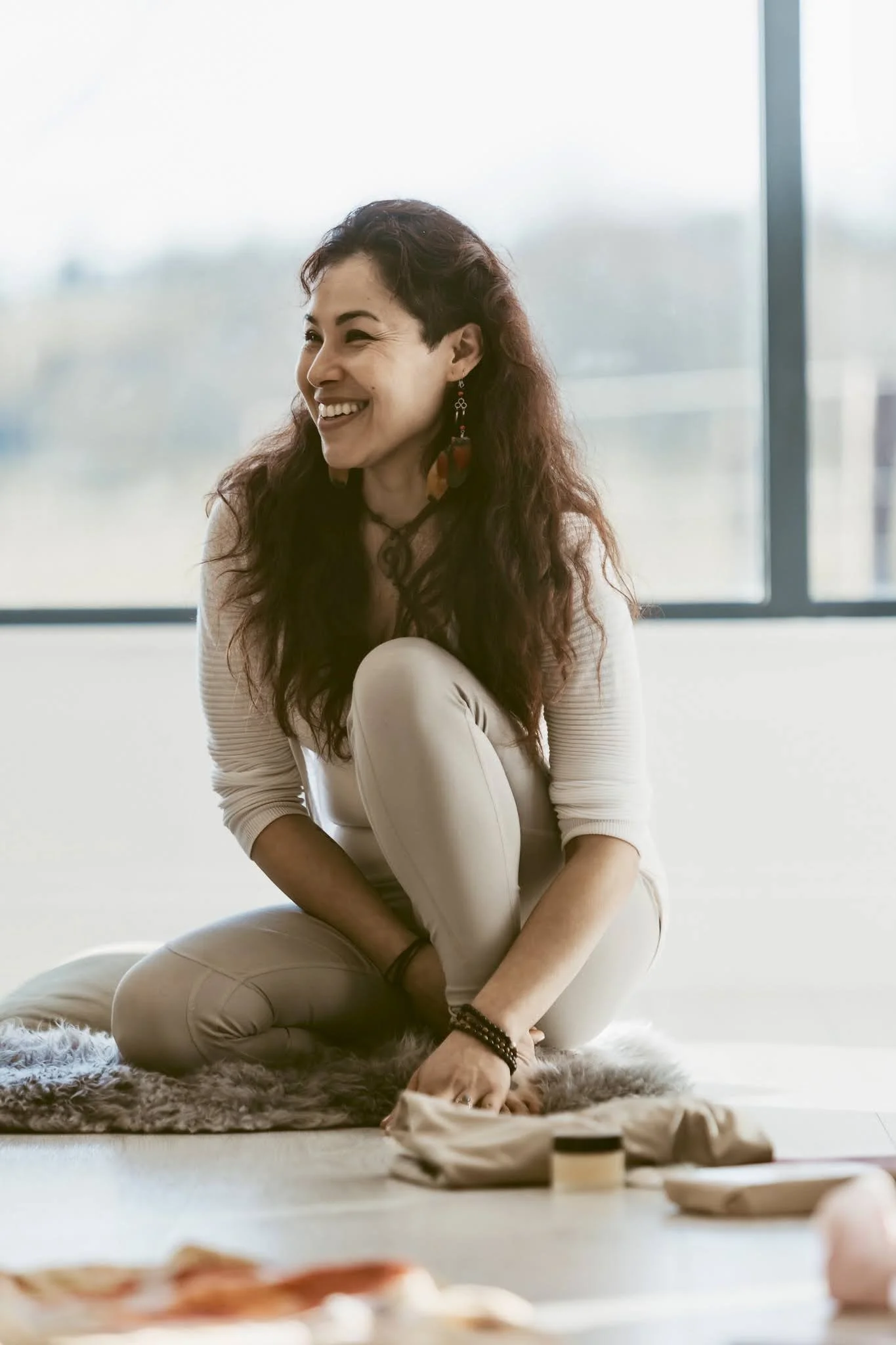 Woman with long dark hair, wearing earrings and casual clothing, sitting on the floor and smiling near a large window.