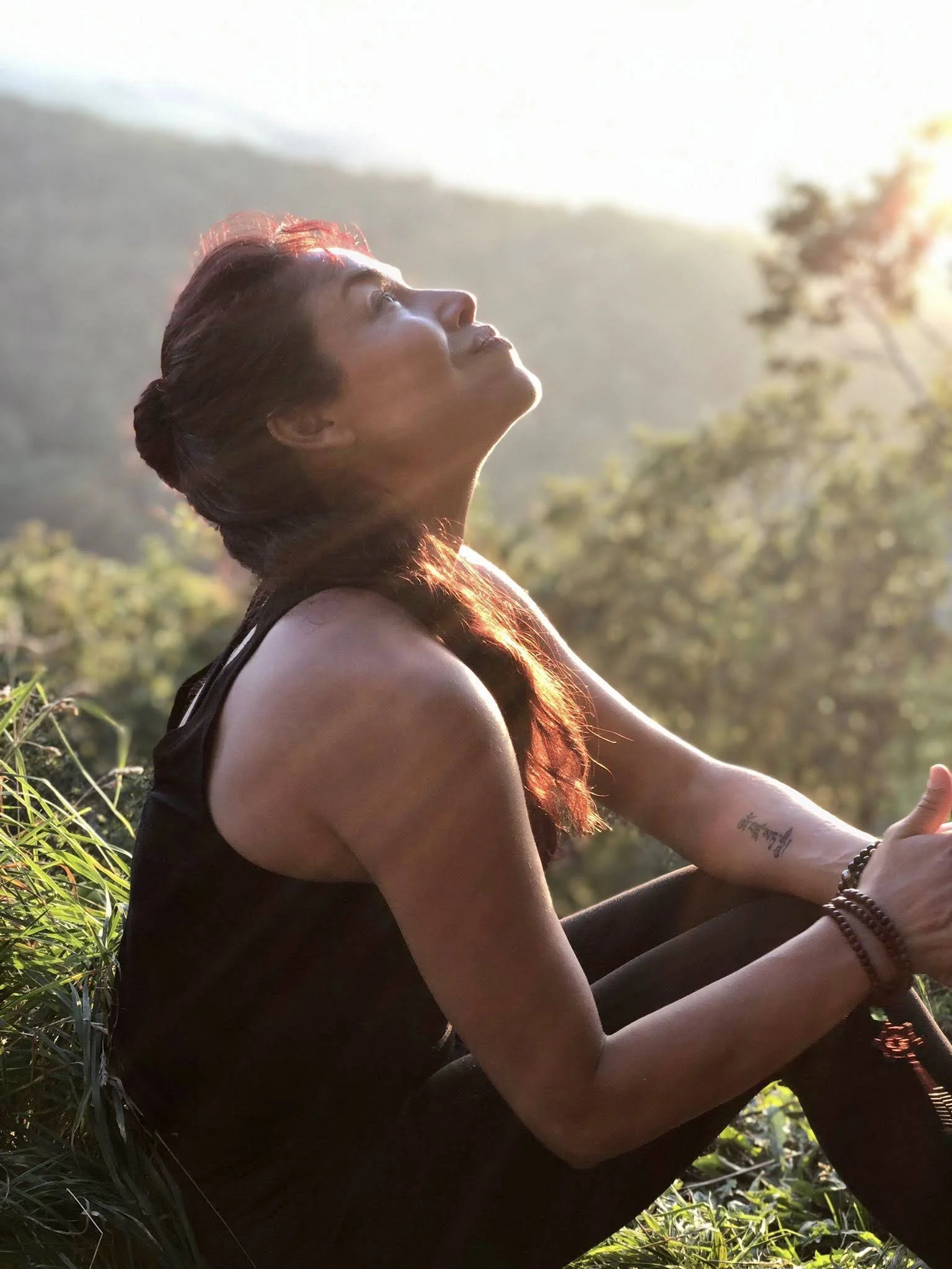 Woman sitting outdoors during sunset, looking up with a peaceful expression, wearing a black sleeveless top and bracelets, with mountains and trees in the background.