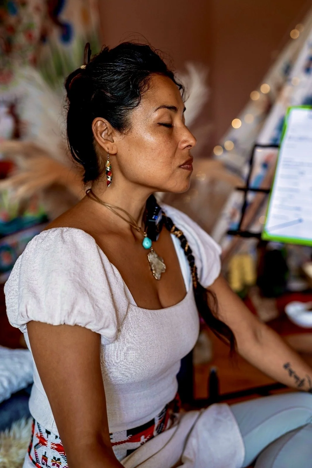 A woman with her eyes closed, sitting peacefully in a relaxed pose, wearing traditional jewelry and attire, with a background of colorful decorations and fairy lights.