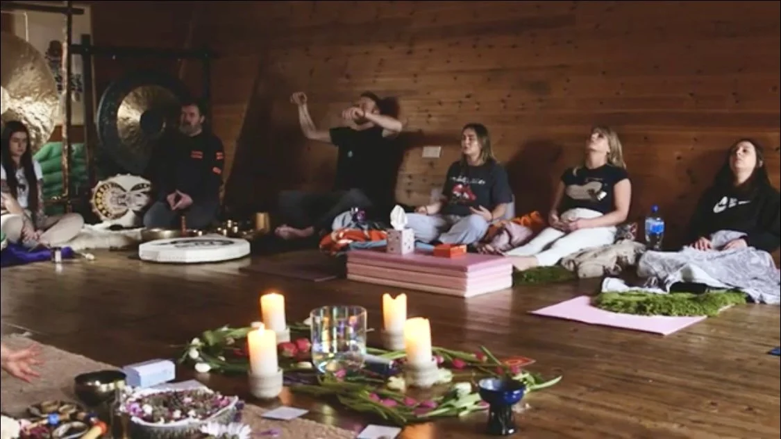 People sitting cross-legged on mats in a wooden room, participating in a meditation or yoga session, with candles, flowers, and spiritual objects in the foreground.