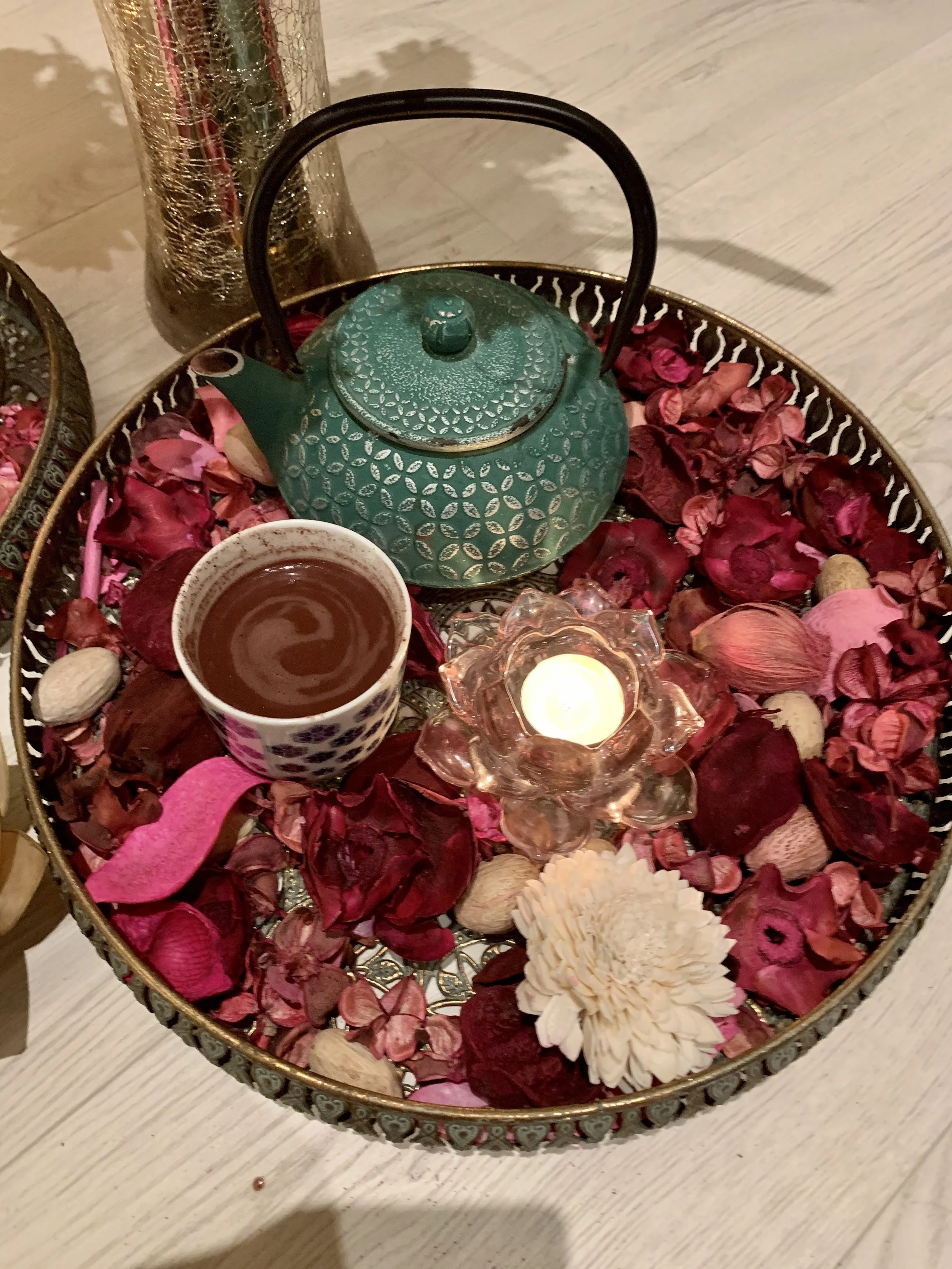 A decorative tray with dried rose petals, nuts, a lit candle, a cup of tea, and a green teapot.