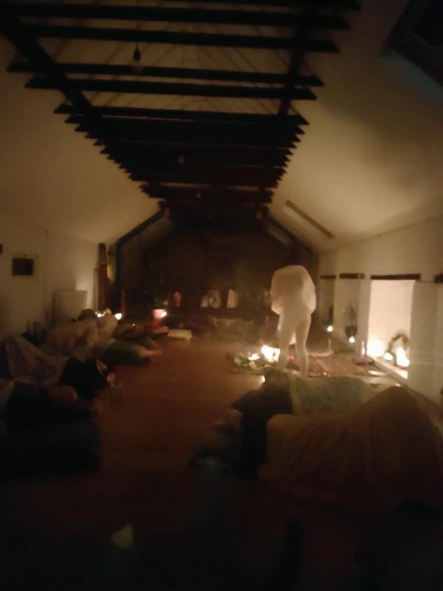 People practicing yoga or meditation in a cozy, candle-lit living room with a high vaulted ceiling and wood beams.