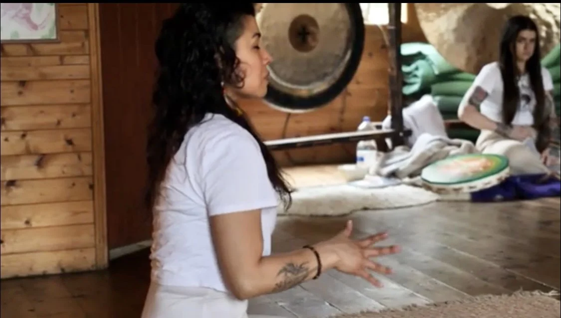 A woman with curly black hair, wearing a white t-shirt, sitting on the floor inside a wooden cabin, with her hands clasped together, and tattoos on her left forearm.