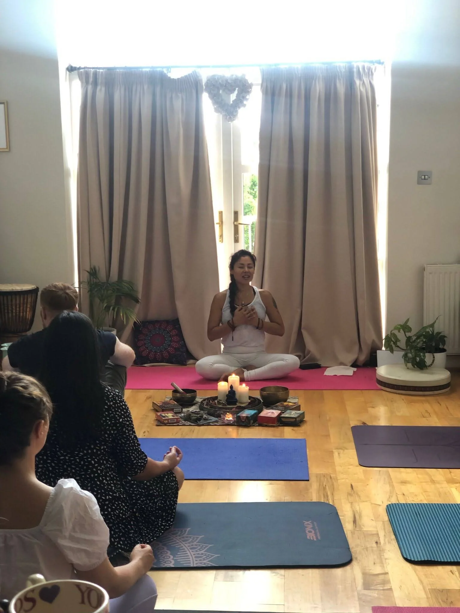 Woman leading a yoga or meditation session in a bright room with large cream curtains, candles, and tarot cards on the floor, with a group of women seated on yoga mats.