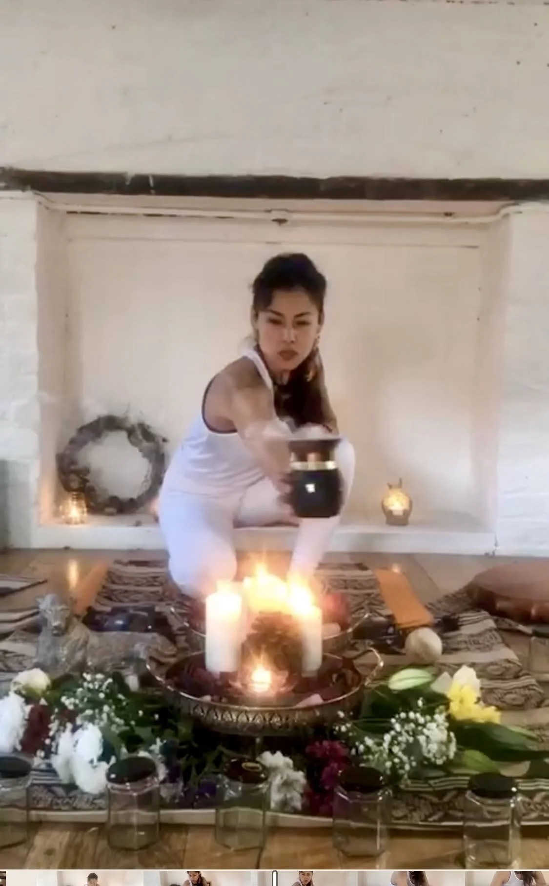 Woman in white crouching on a decorated mat, reaching toward candles and flowers in a ritual setting.