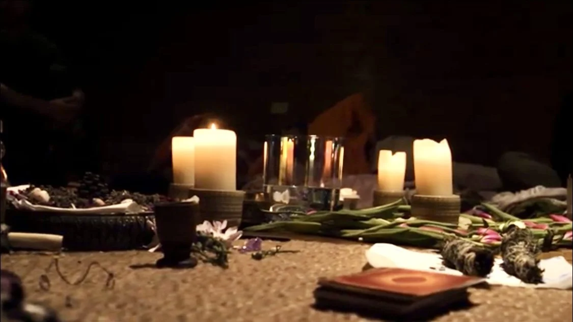 Candles, flowers, and objects on a surface in a dimly lit setting, possibly for a memorial or ritual.