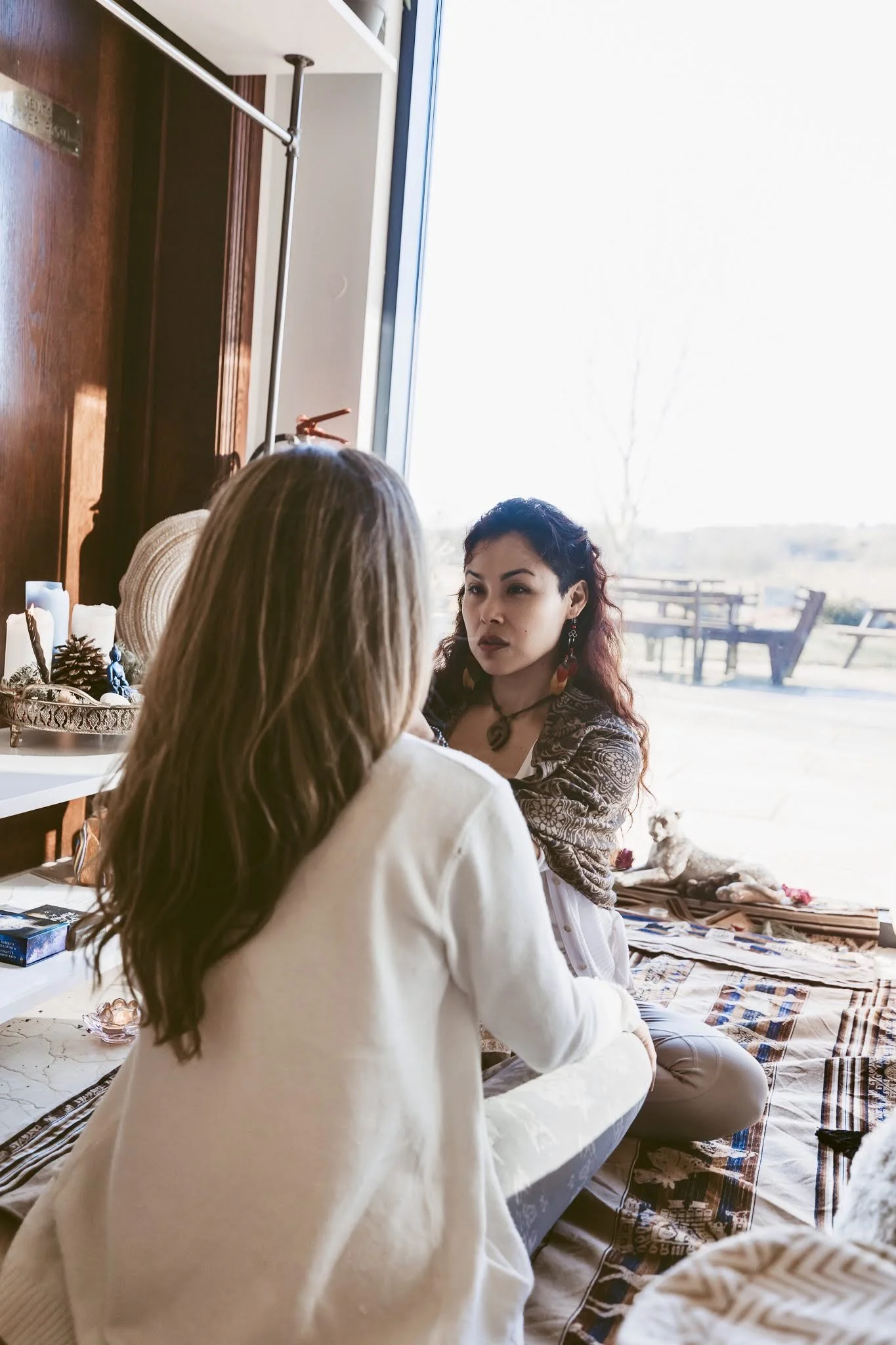 Two women sitting on a blanket inside a room, engaging in a conversation. One woman has long blonde hair, and the other has dark hair with red highlights, wearing jewelry and a patterned shawl. There are decorative items and a dog outside on the pati