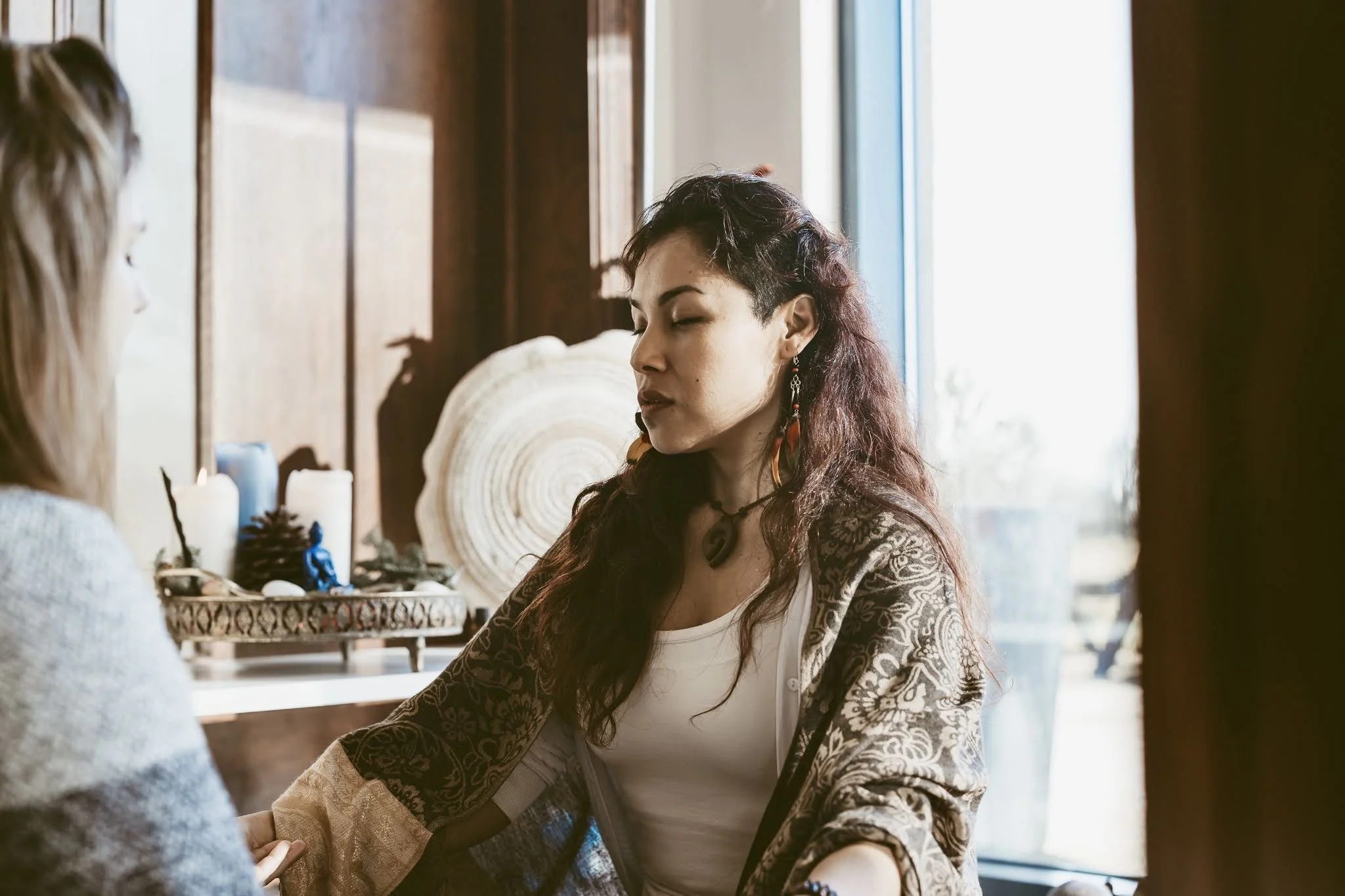 A woman with long curly hair, wearing earrings, a necklace, and a patterned jacket, appears to be meditating or practicing energy work with her eyes closed, sitting across from another woman in a well-lit room with candles and decorative objects in t