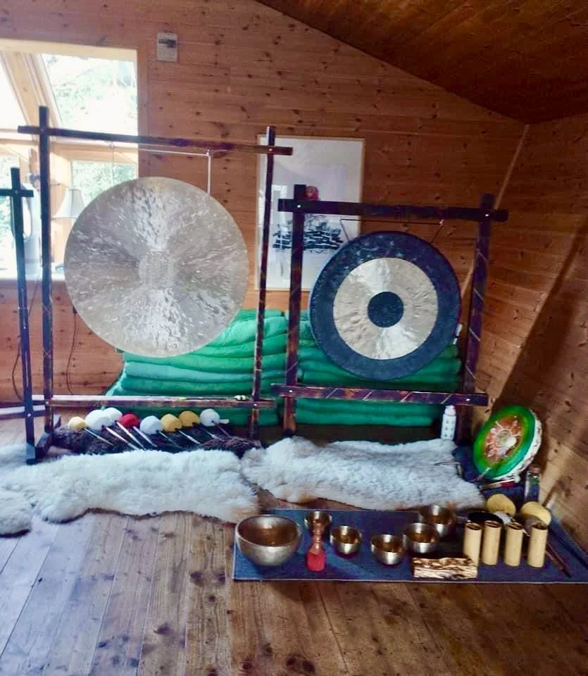 Room with wooden walls and floor, two large gongs, an assortment of singing bowls, shields, and mallets, with sheepskin rugs on the floor and a window in the background.