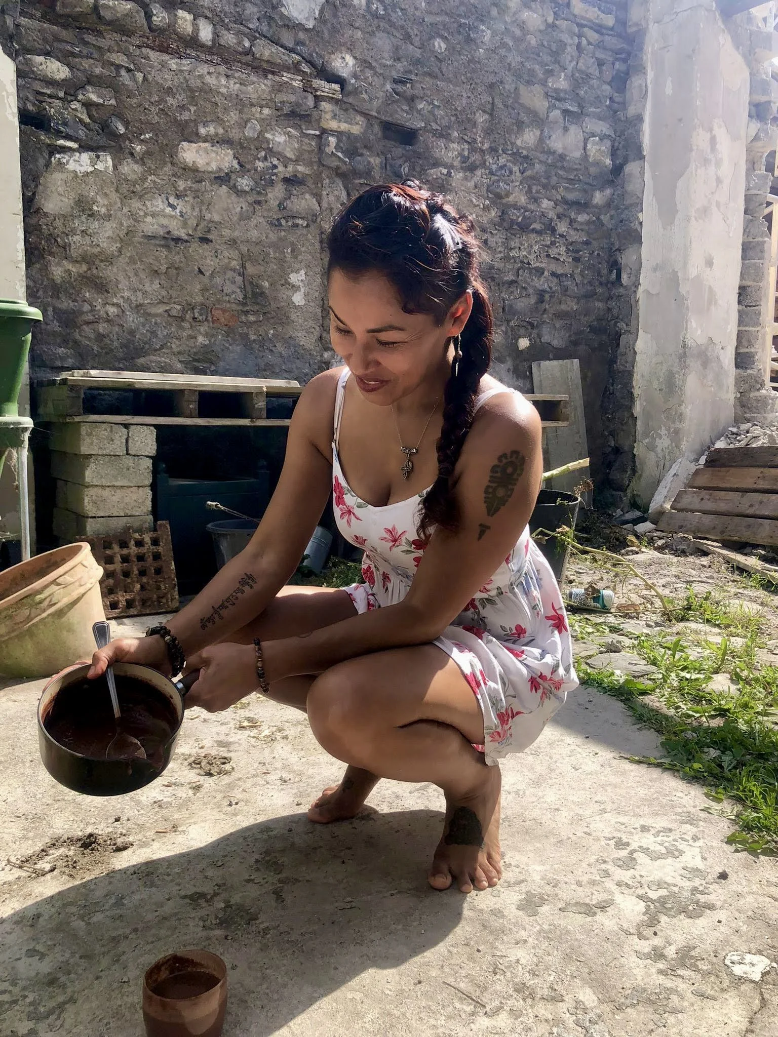 A woman in a white floral dress crouching outdoors, holding a dark bowl with a spoon, smiling, with a stone wall and plants in the background.