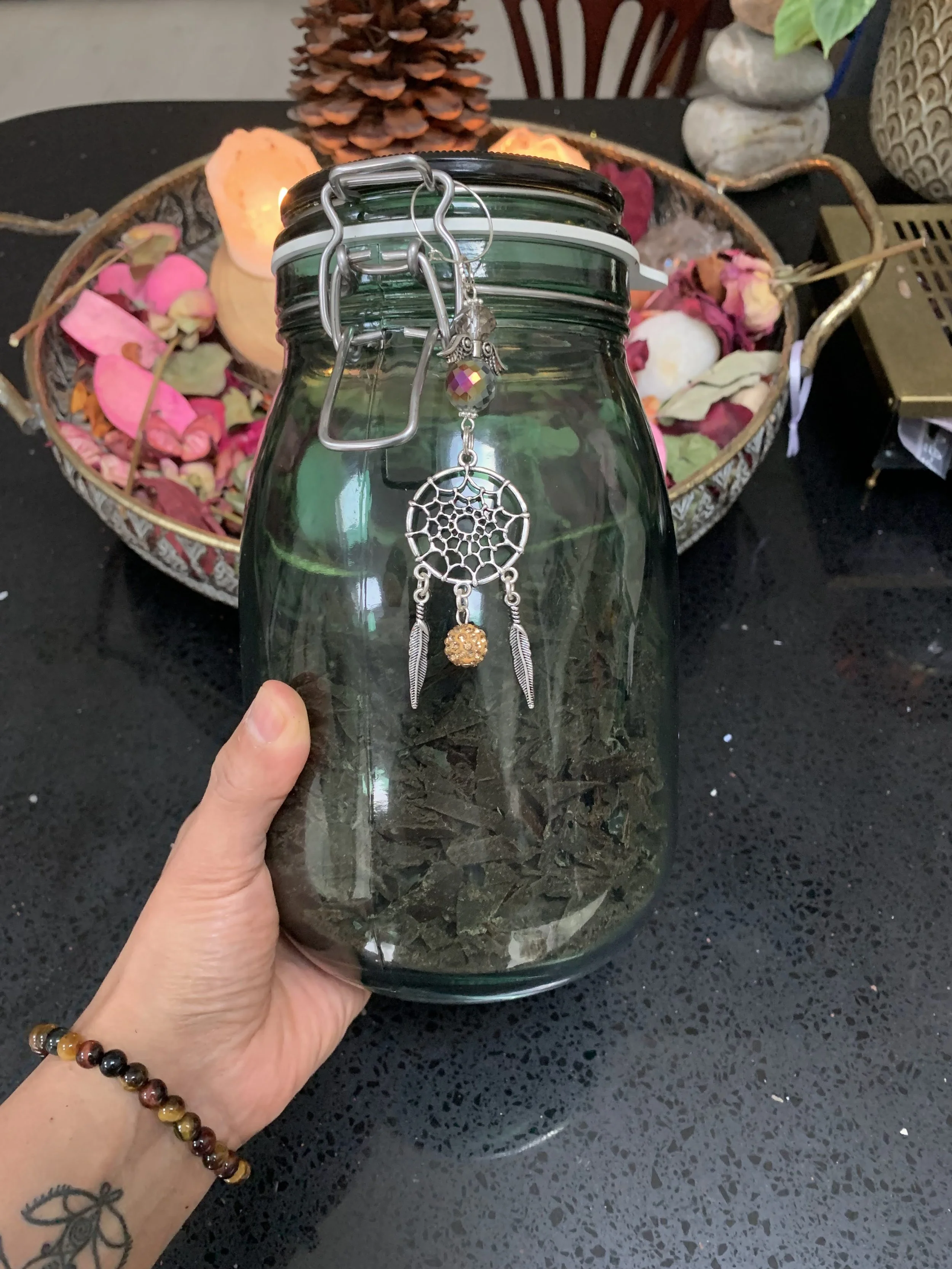 A person's hand holding a large glass jar filled with dried herbs or leaves, with jewelry including a dreamcatcher pendant hanging from the jar's lid. In the background, a decorative basket contains colorful dried flower petals and other decorative i