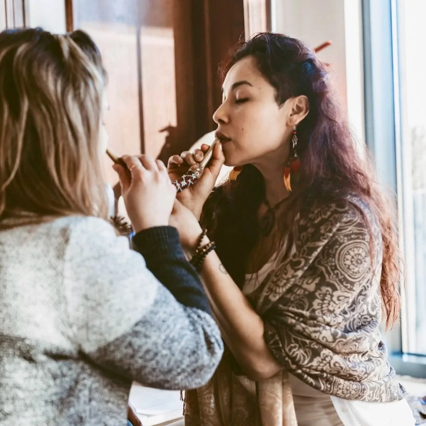 Two women, one with long curly hair and the other with short dark hair, are smoking cigarettes together inside a room with wooden walls and a window.