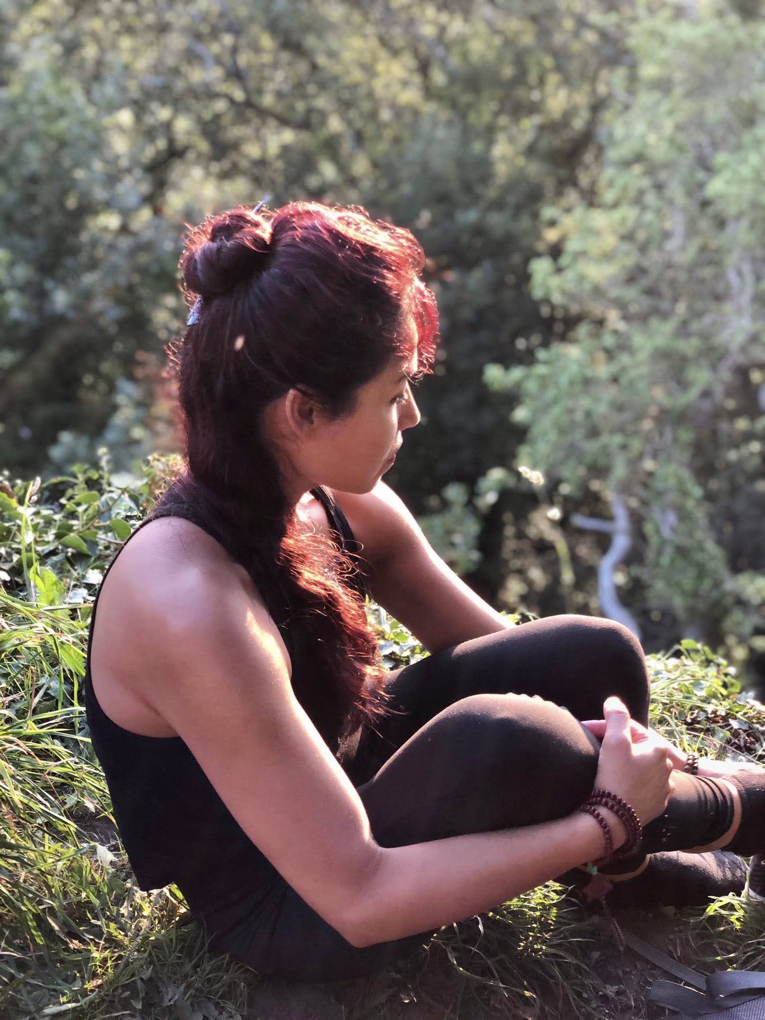 A woman sitting on the ground in a natural outdoor setting, illuminated by warm sunlight, with her knees pulled close and hair styled in a bun, looking towards the right.