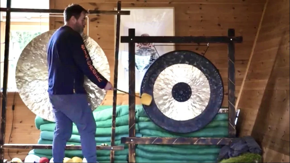 A man is striking a gong with a mallet inside a wooden room with green blankets or mats stacked against the wall.