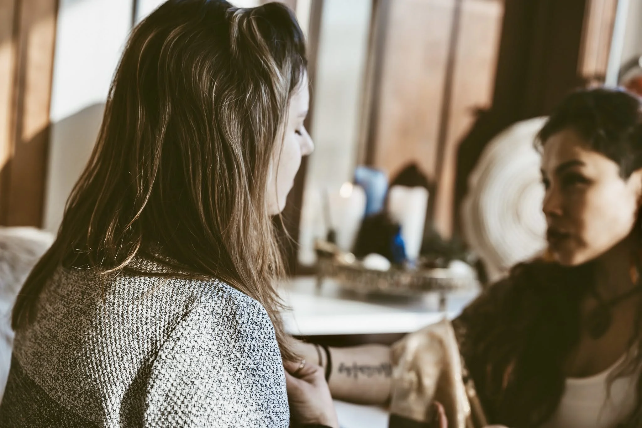 A woman with brown hair looking down, and a tattoo on her arm, sitting across from a woman with short dark hair and tattoos, in a cozy, wooden-paneled room.