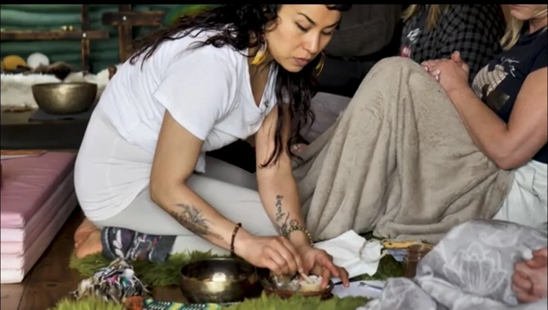 Woman with long curly hair, tattoos, and colorful earrings performing a ritual or prayer with others in a spiritual or ceremonial setting.