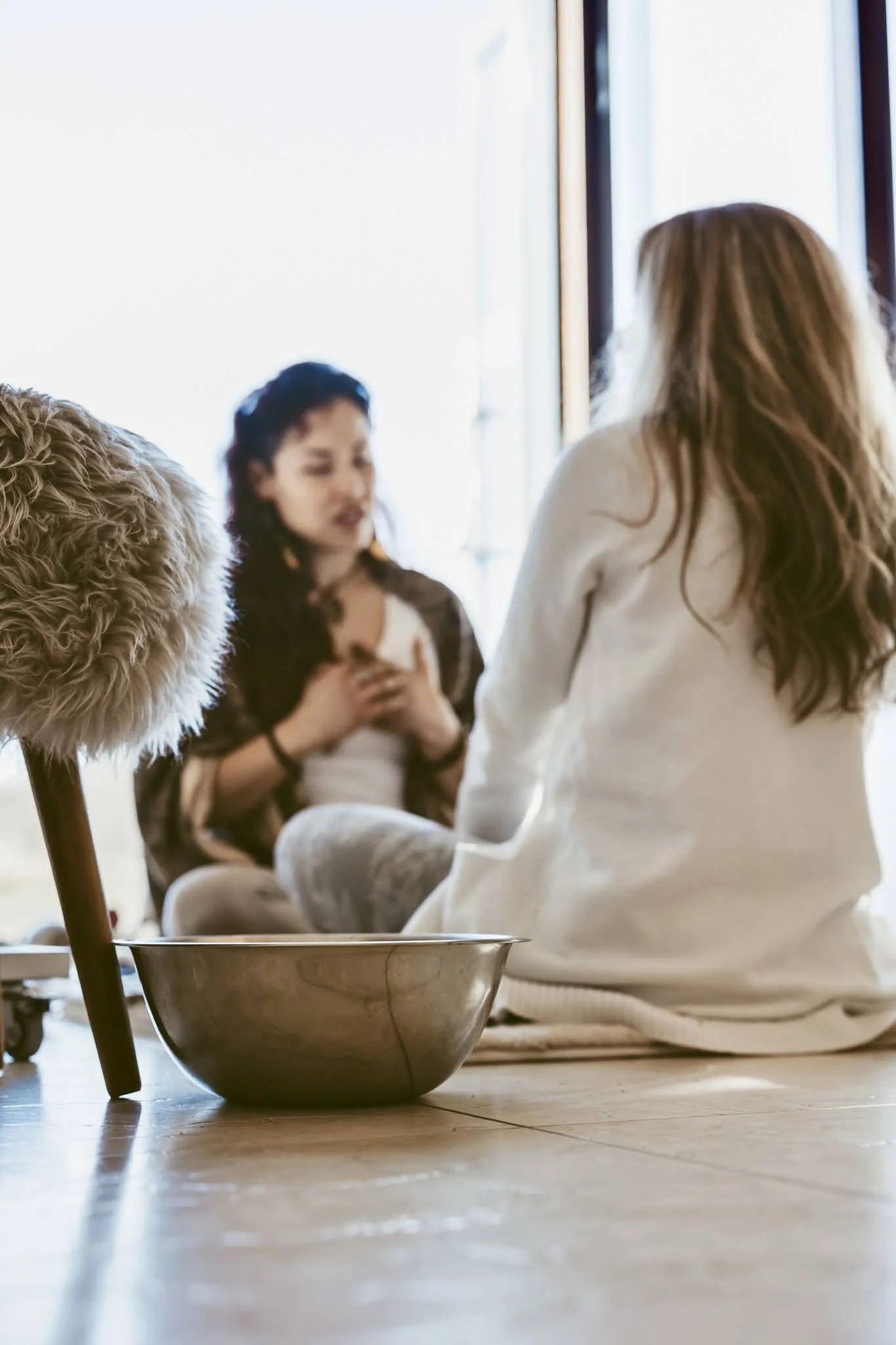 Two women sitting on the floor indoors, one with long hair facing away, the other with dark hair holding her chest, near a window with natural light, and a metal bowl and fluffy object in the foreground.