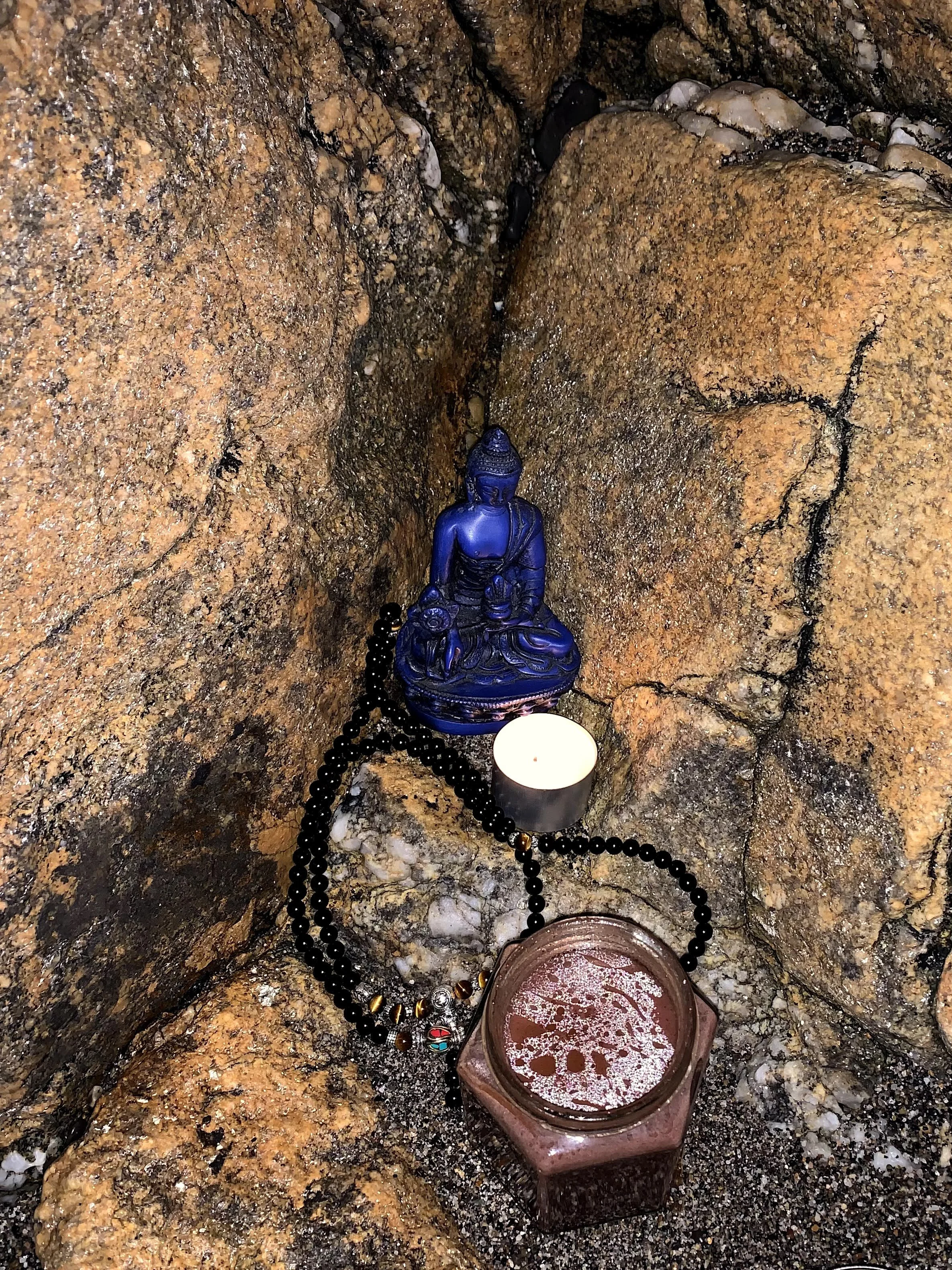 A small altar with a blue Buddha statue, a lit tealight candle, a beaded necklace with gold accents, and a pinkish jar of liquid, set among rocks.