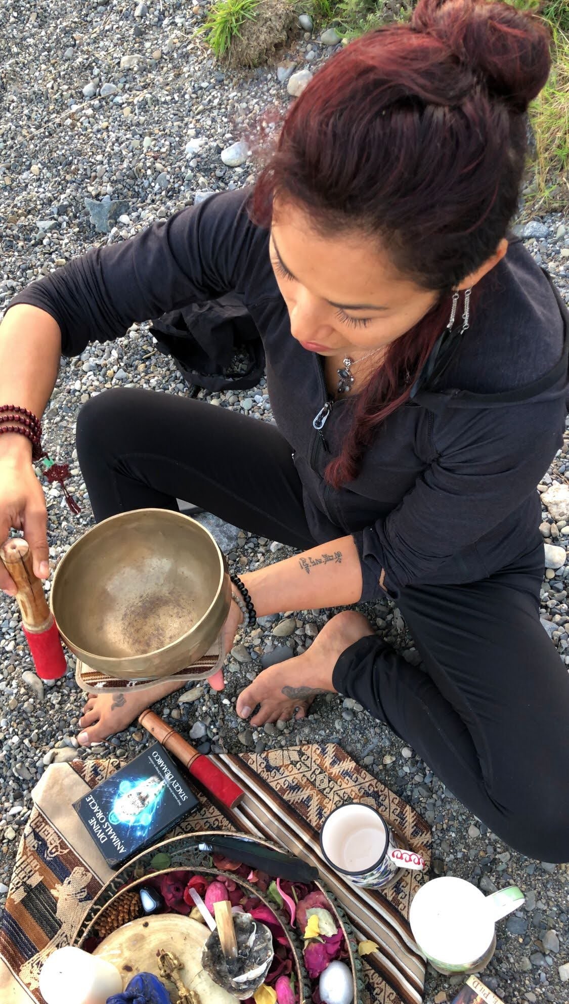 A woman with red hair, dressed in black, sits on a rocky surface, holding a singing bowl. She has a tattoo on her forearm and wears beaded jewelry. Surrounding her are various objects including a book titled 'Silent Wisdom', a mug, a glass, and spiri