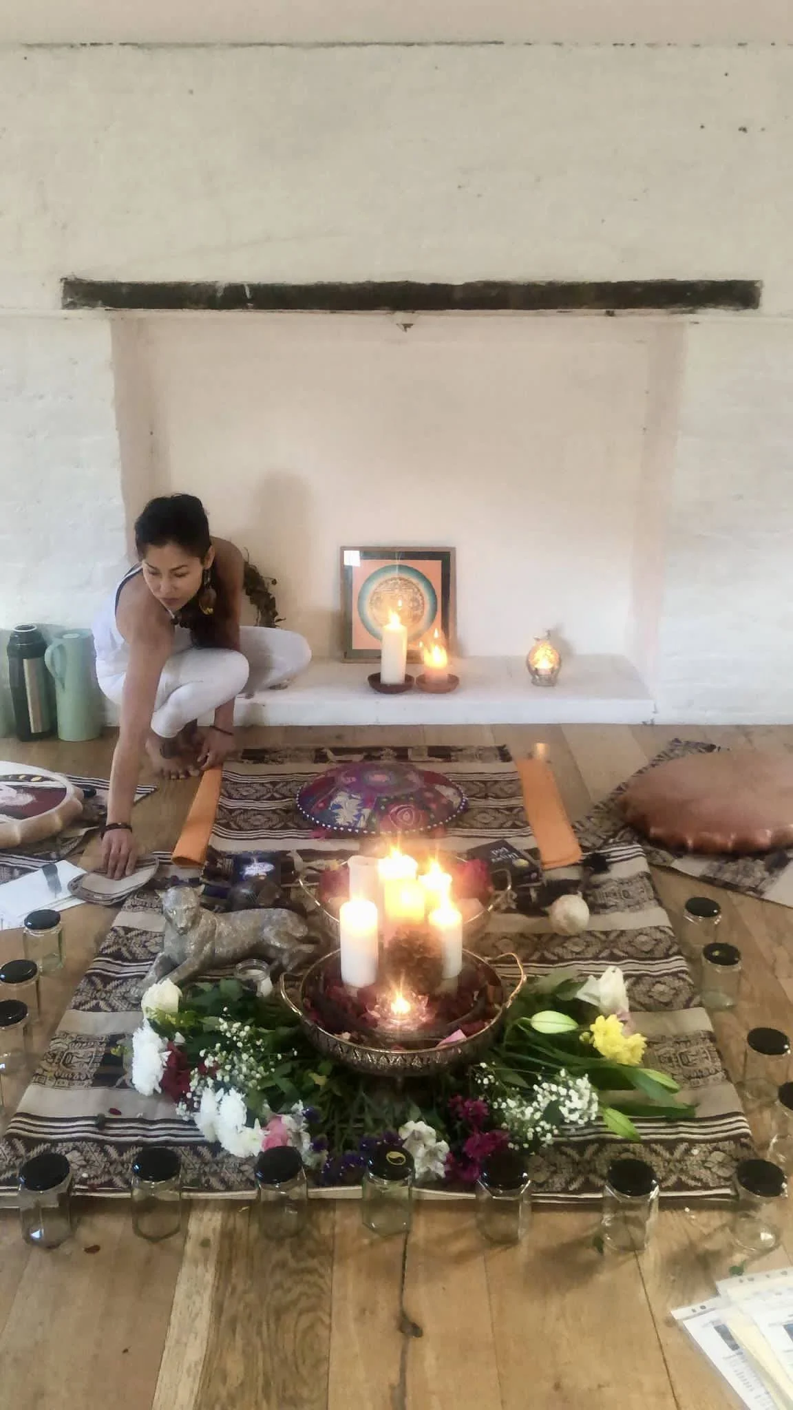 A woman participating in a spiritual or meditative practice, sitting on a patterned blanket surrounded by candles, flowers, jars, and spiritual artifacts on a wooden floor in front of a white fireplace.