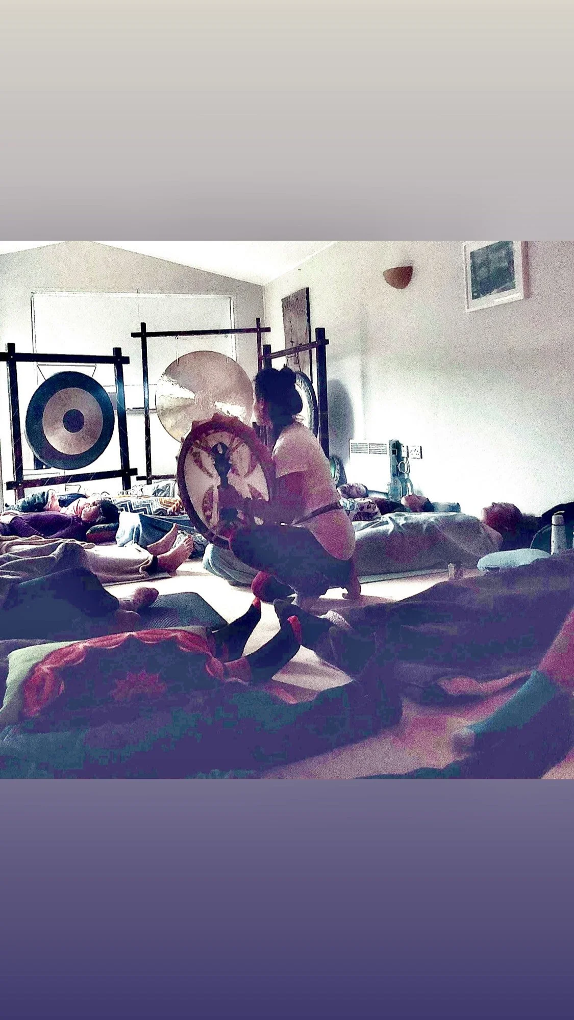 A woman is sitting cross-legged on a platform, holding a shield, during a yoga session in a bright room with multiple people lying on mats in different poses.