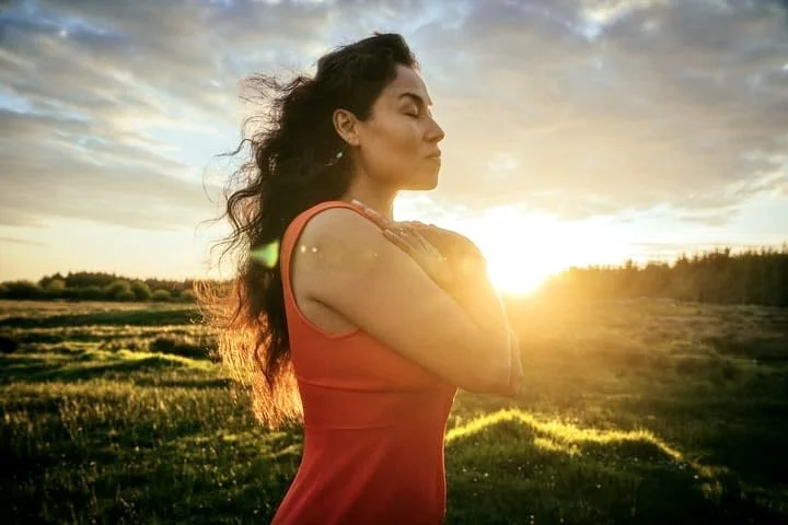 A woman meditating outdoors during sunrise or sunset with her eyes closed and hands on her chest, in a grassy field.