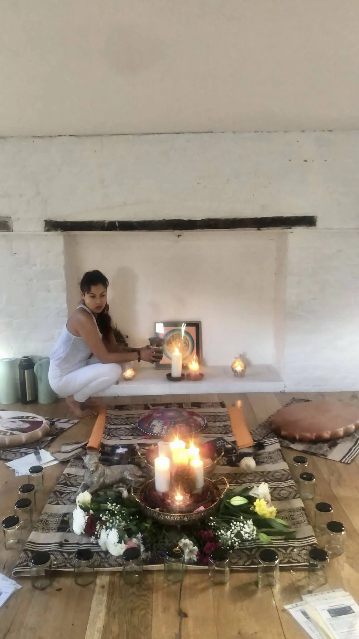 A woman in white pajamas is sitting on a rug in front of a fireplace, lighting candles surrounded by flowers, glass jars, and candles, with cushions and photographs nearby.
