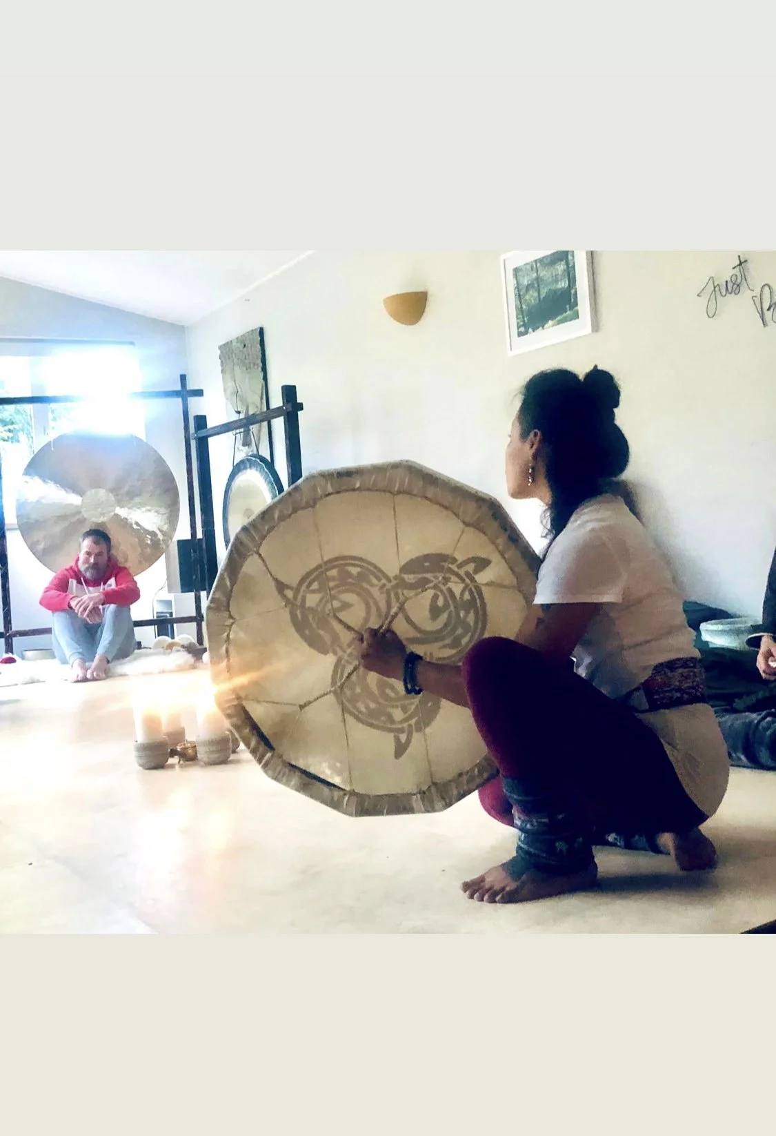 A woman sitting on the floor with a tattooed drum, with a light-skinned man sitting cross-legged near large gongs in a room with natural light, candles, and wall art.
