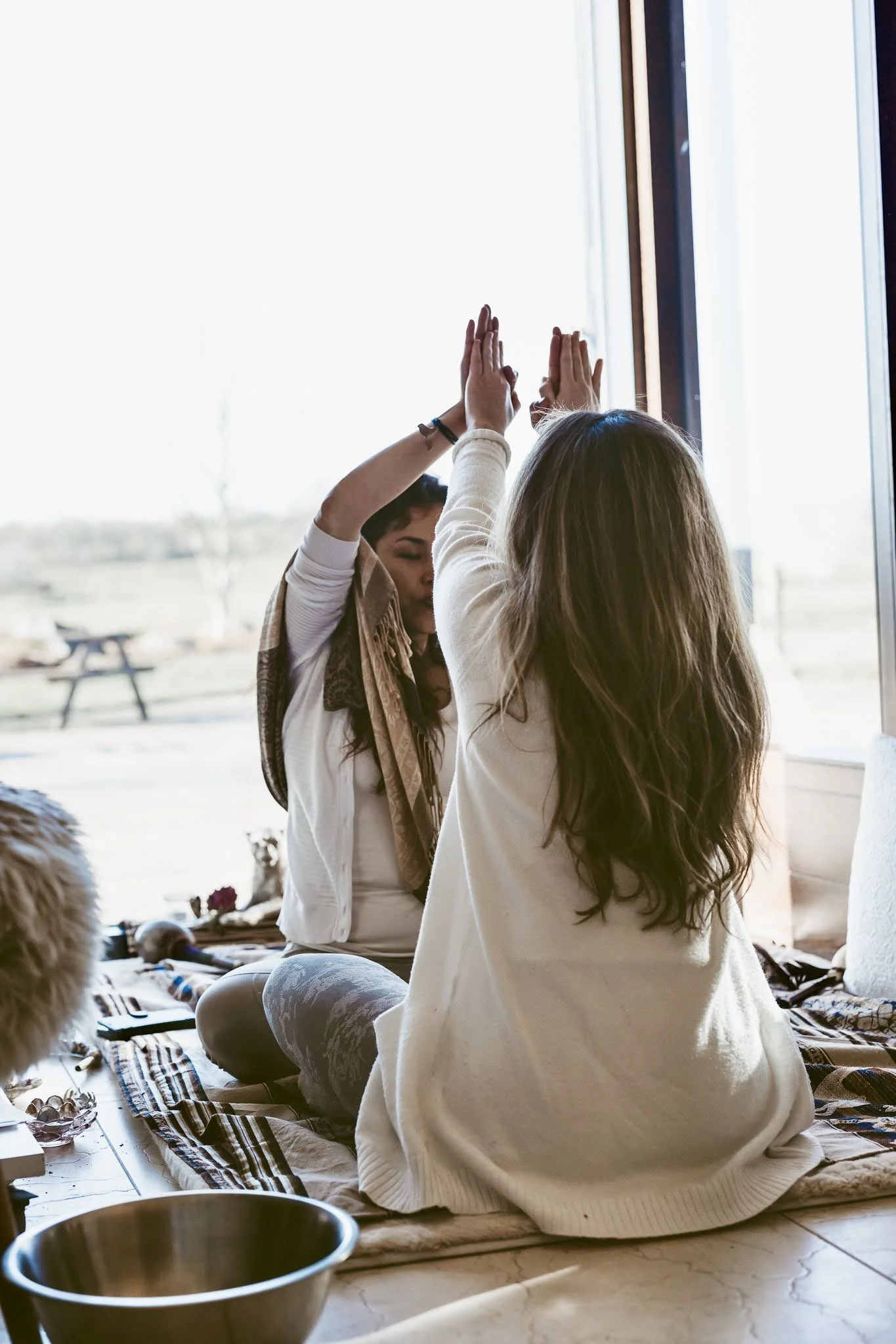 Two women practicing yoga together indoors near a large window, sitting on a rug with various items around them.