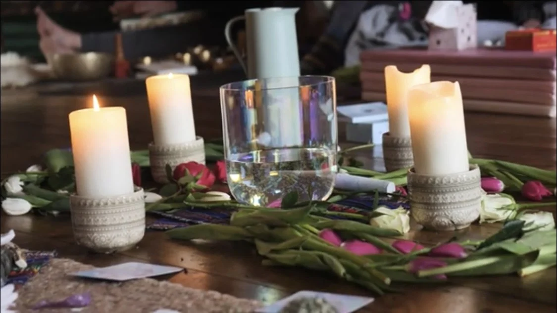 Table decorated with candles, flowers, and a glass of water, surrounded by books and miscellaneous items