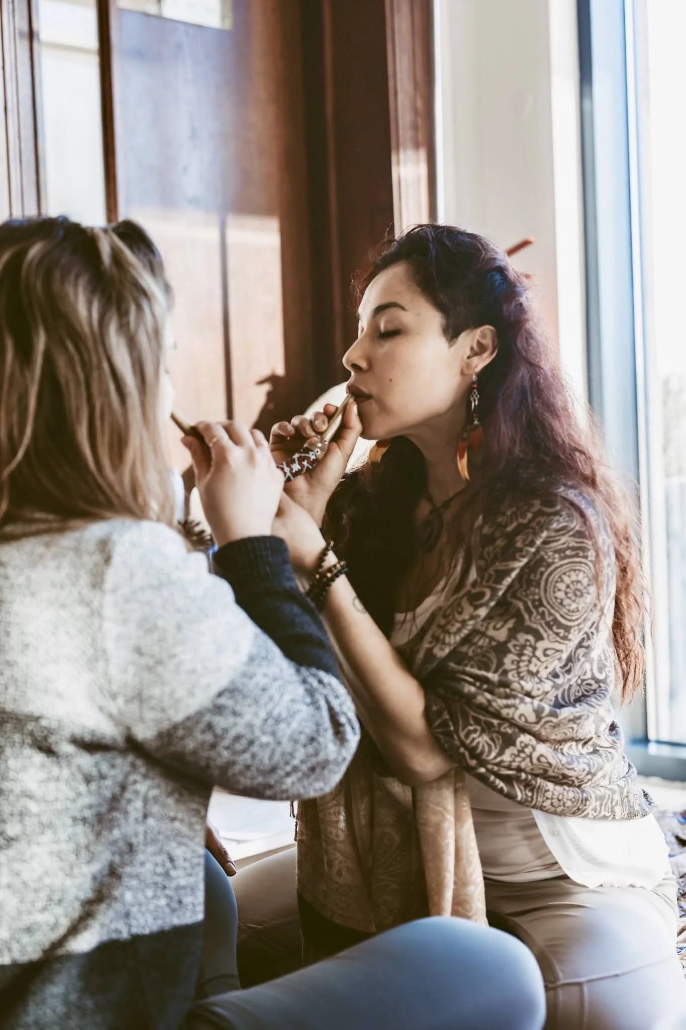A woman with dark, curly hair and expressive earrings has her lips painted by another woman with blonde hair, inside a room with large windows and warm lighting.