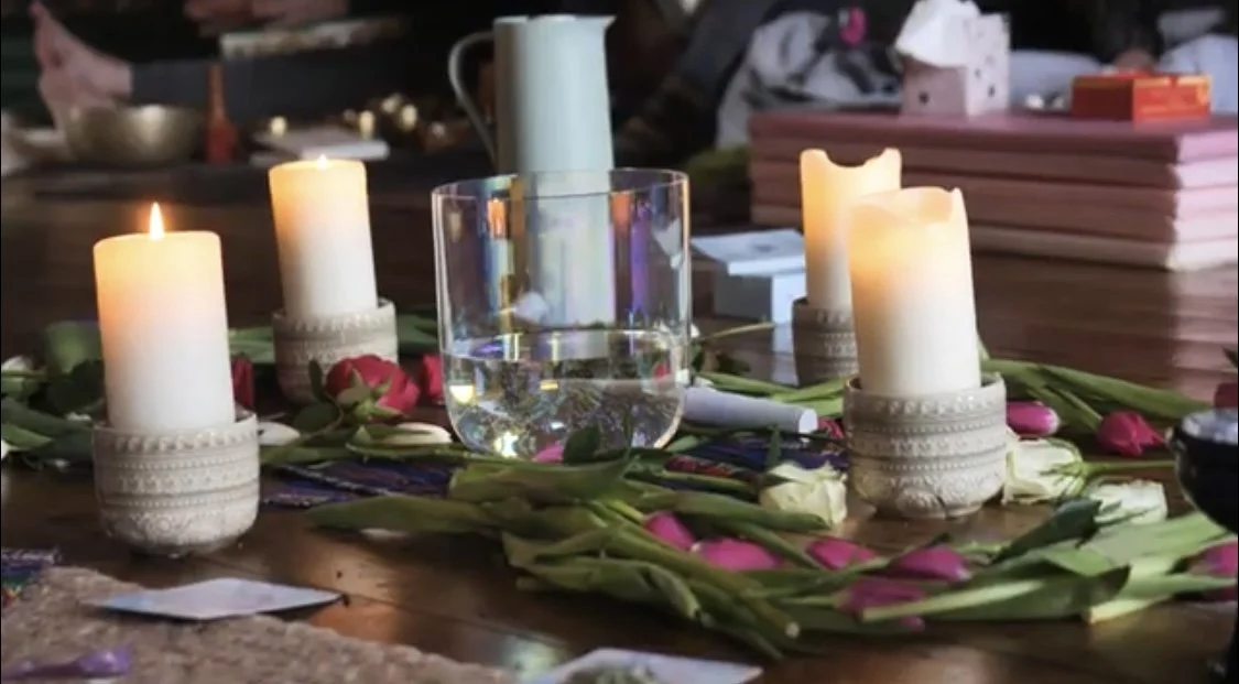 Table with four lit candles, a glass of water, pink and white flowers arranged in a circle, and various objects in the background.