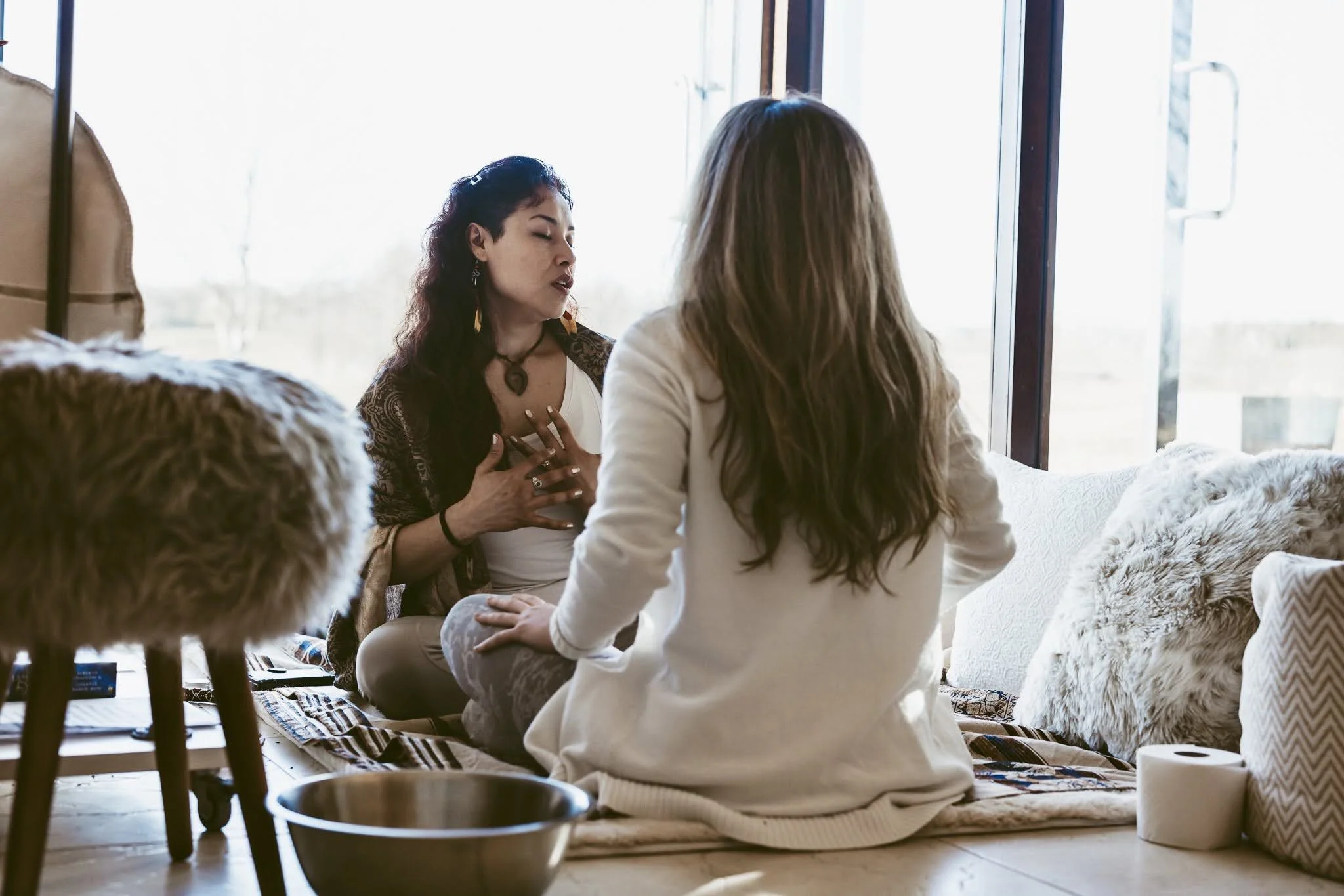 Two women are sitting on a padded mat by a large window, engaging in a heartfelt conversation. One woman is touching her chest, eyes closed, while the other listens intently. The room is cozy with furry pillows and a roll of toilet paper nearby.